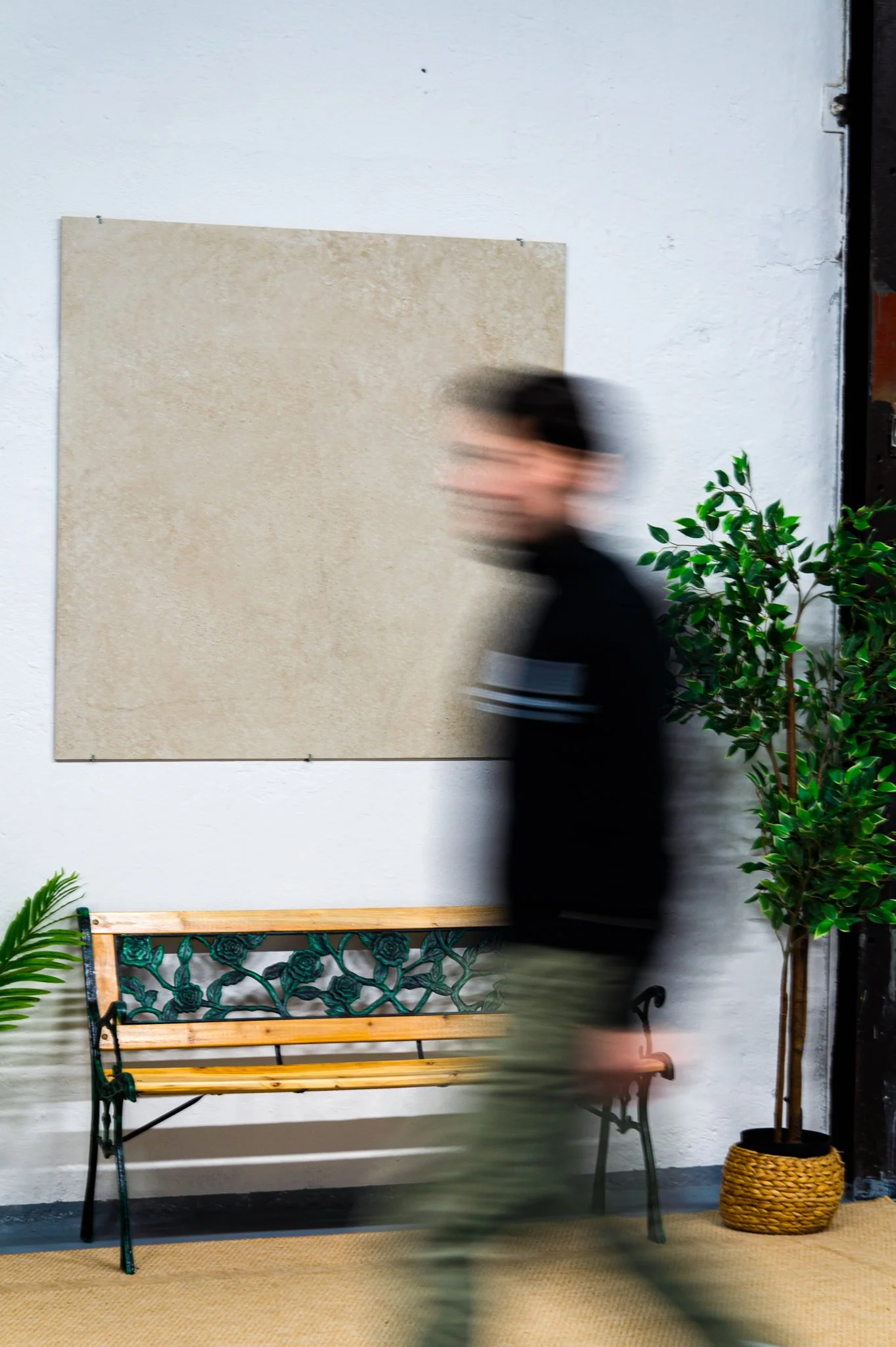 Eine Person geht vorbei an einer weißen Wand mit einem beigefarbenen Bild und sitzt auf einer schwarzen Metallbank mit Holzlatten. Es gibt zwei grüne Pflanzen, eine in einem Korb und eine andere in einem Topf, im Raum. Aufgenommen von Hilal Studios.