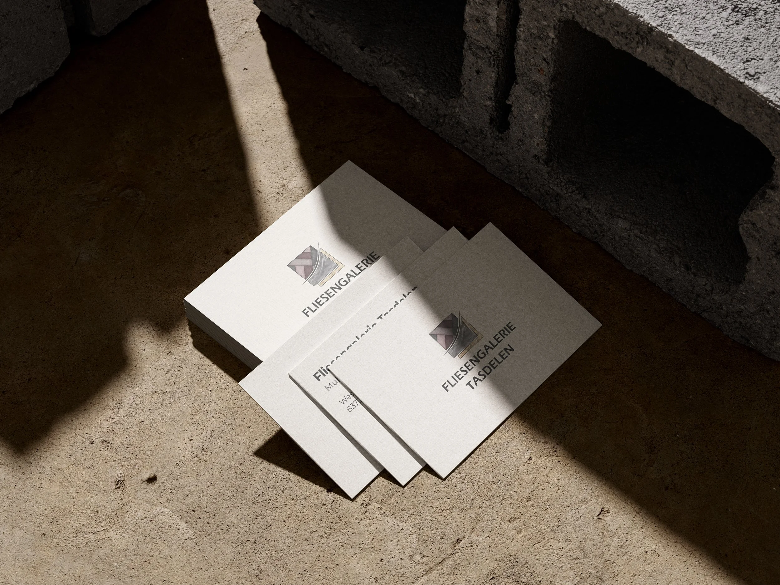 Drei weiße Flyer mit dem Logo einer Fliesengalerie und dem Text "Fliesengalerie Tasdelen" liegen auf einem rohen Betonboden, mit Schatten und einer Ziegelmauer im Hintergrund. Erstellt von Hilal Studios.