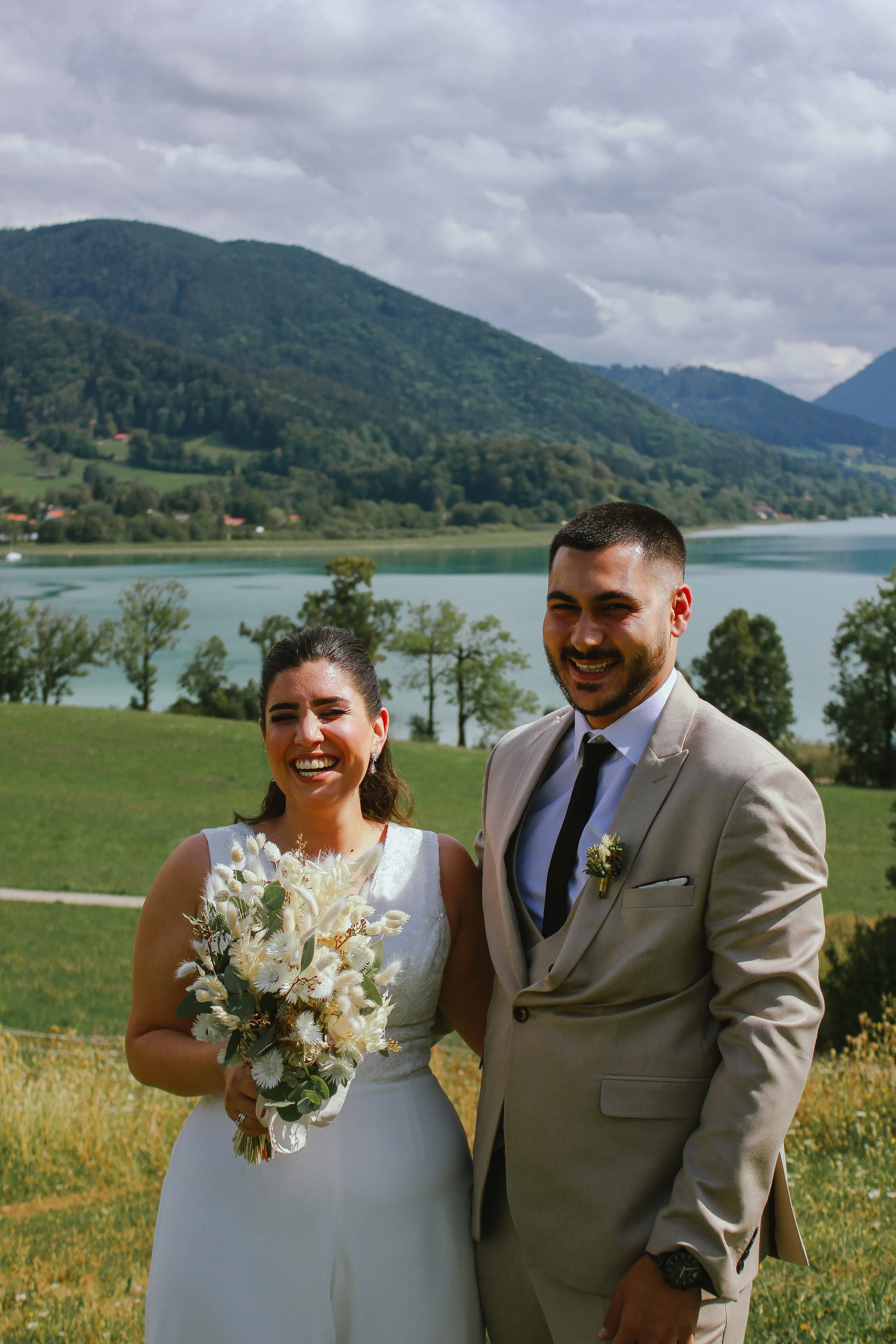 Ein glückliches Brautpaar bei ihrer Hochzeit im Freien, mit Bergen, See und grüner Wiese im Hintergrund. Aufgenommen von Hilal Studios.