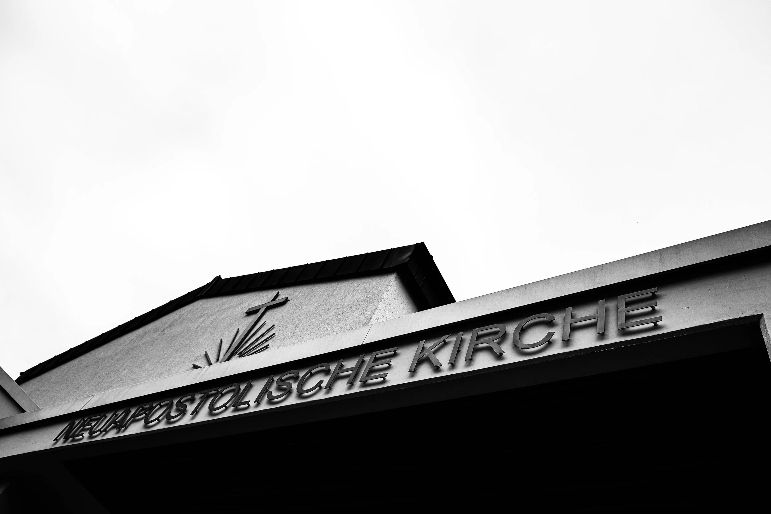 Schwarz-weiß-Foto der Vorderseite einer Kirche mit Schriftzug 'Neuperlachische Kirche' und einem Uhrturm mit Kreuz, Himmel im Hintergrund. Aufgenommen von Hilal Studios.