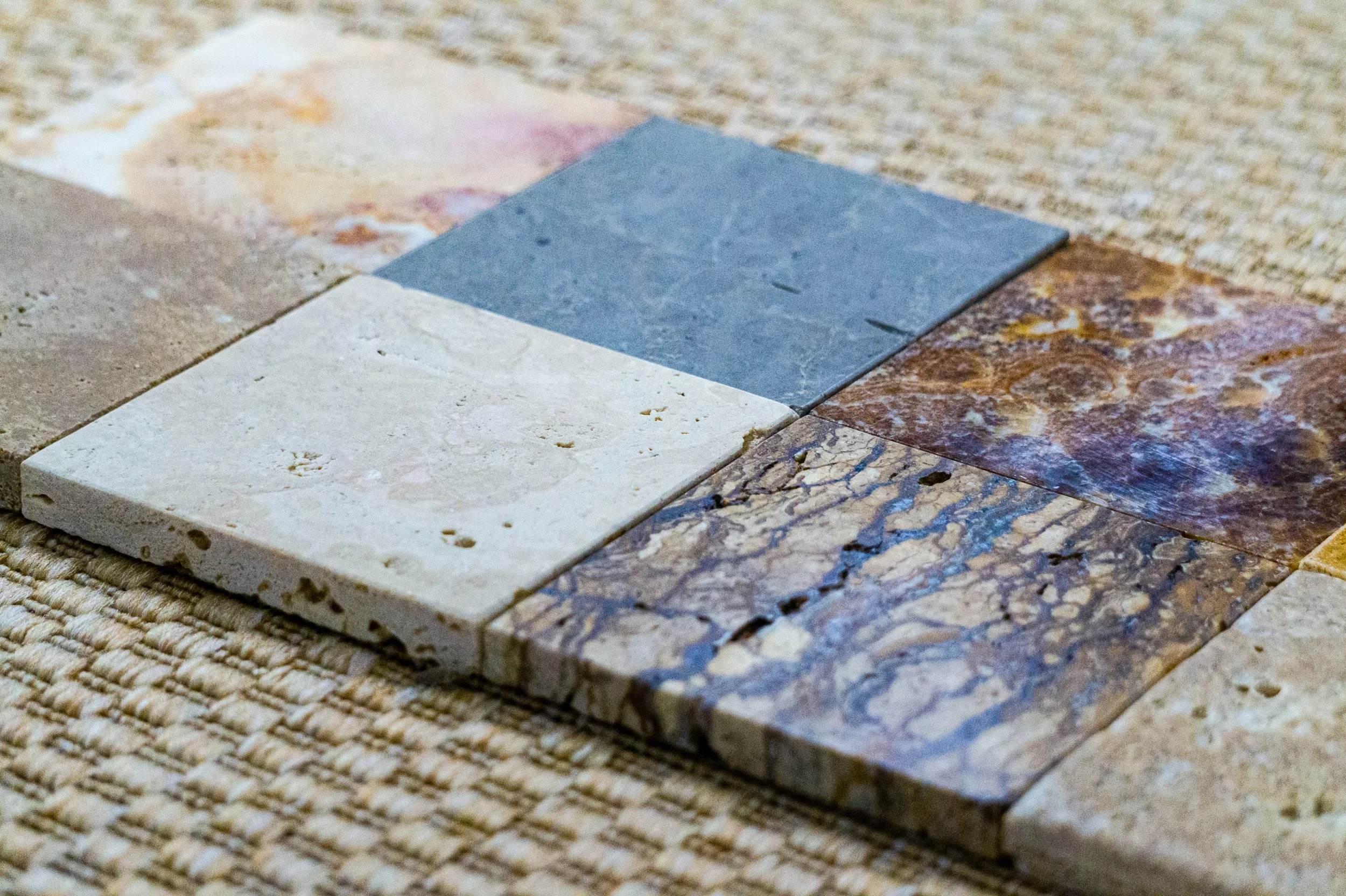 Verschiedene quadratische Natursteinfliesen in unterschiedlichen Farben und Mustern auf einem Teppichboden. Aufgenommen von Hilal Studios.