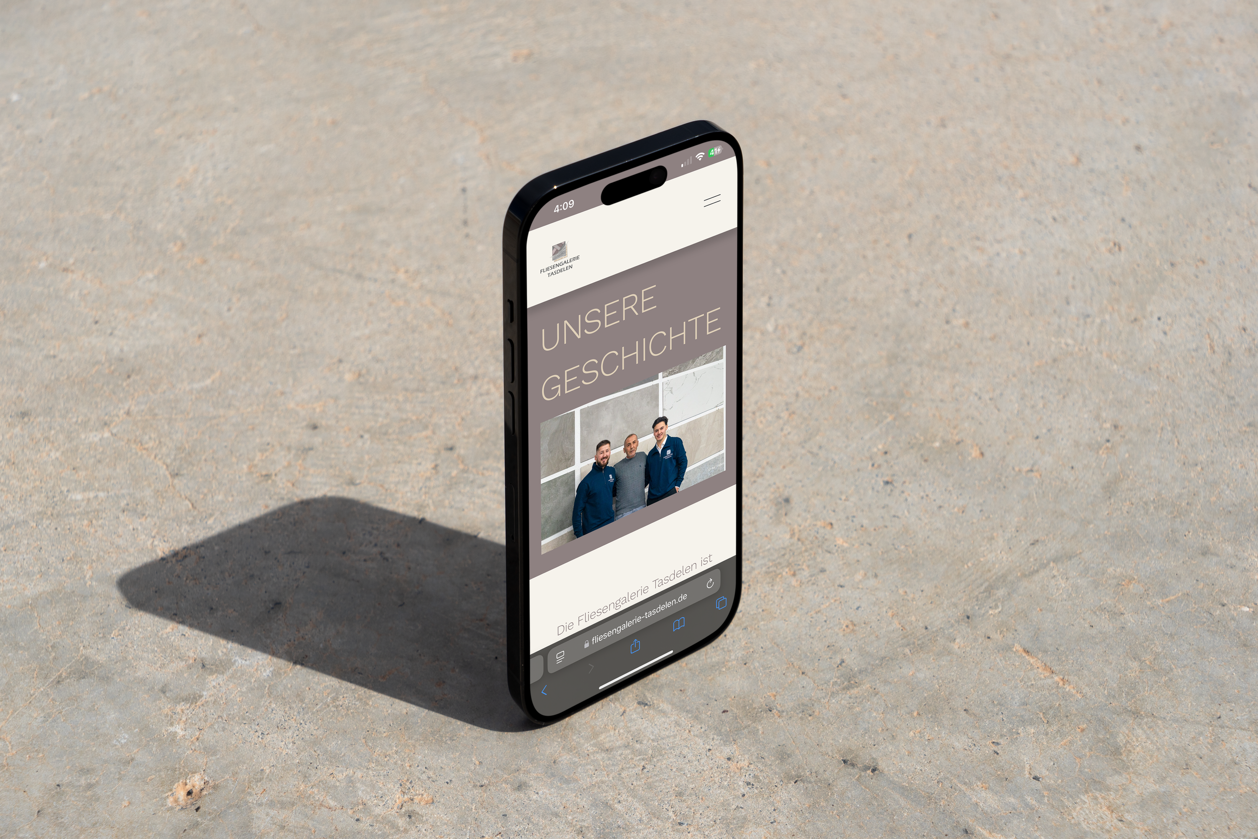 Smartphone auf Sand mit Webseite der Fliesengalerie Tasdelen, Überschrift 'Unsere Geschichte' und Bild von drei Männern. Erstellt von Hilal Studios.