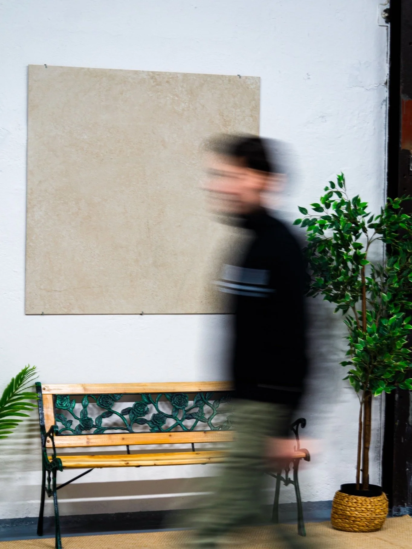 Ein sich bewegender Mann in einem schwarzen Pullover, neben einer grünen Pflanze und einer Gartenbank, vor einer weißen Wand mit einer großen quadratischen Tafel. Aufgenommen von Hilal Studios.