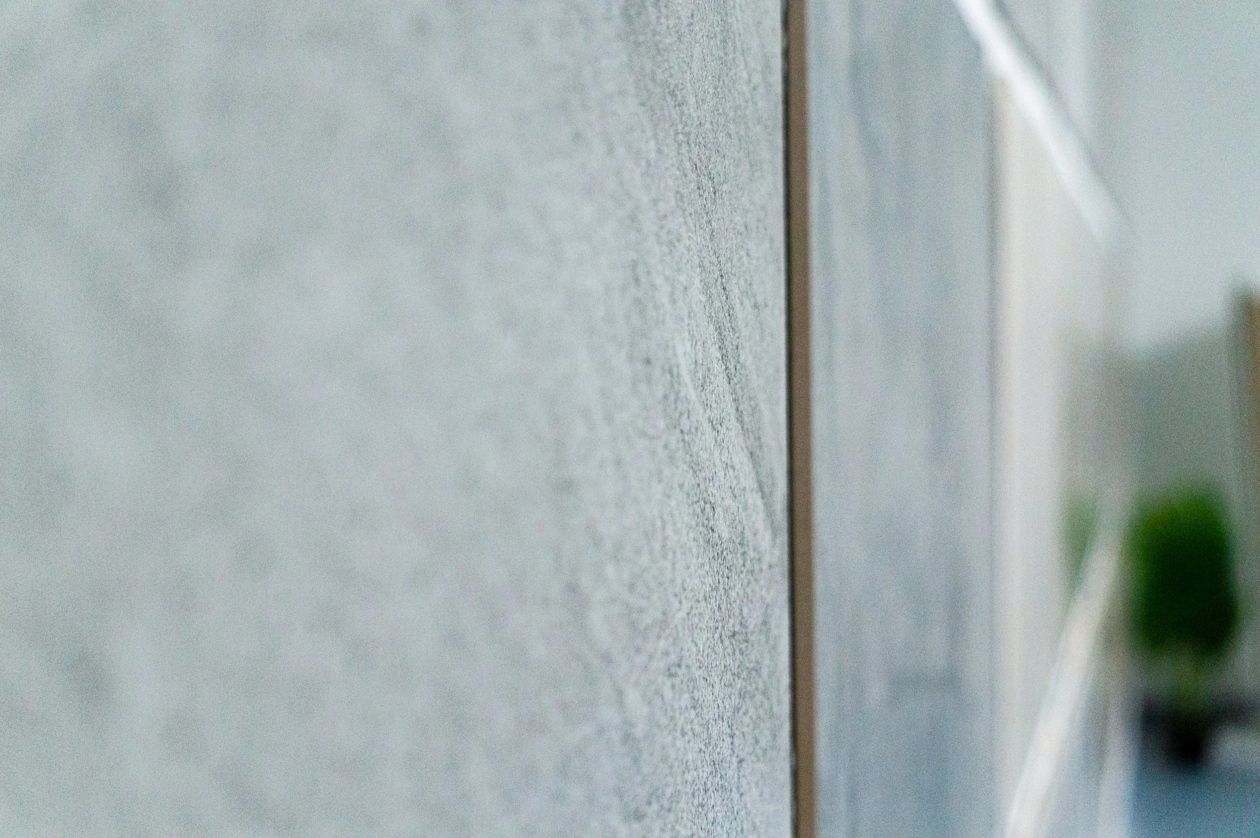 Nahaufnahme einer Wand mit präzisen Fliesen im Innenbereich, die Wand ist hellgrau mit einer Textur, im Hintergrund ist eine kleine Grünpflanze sichtbar. Aufgenommen von Hilal Studios.