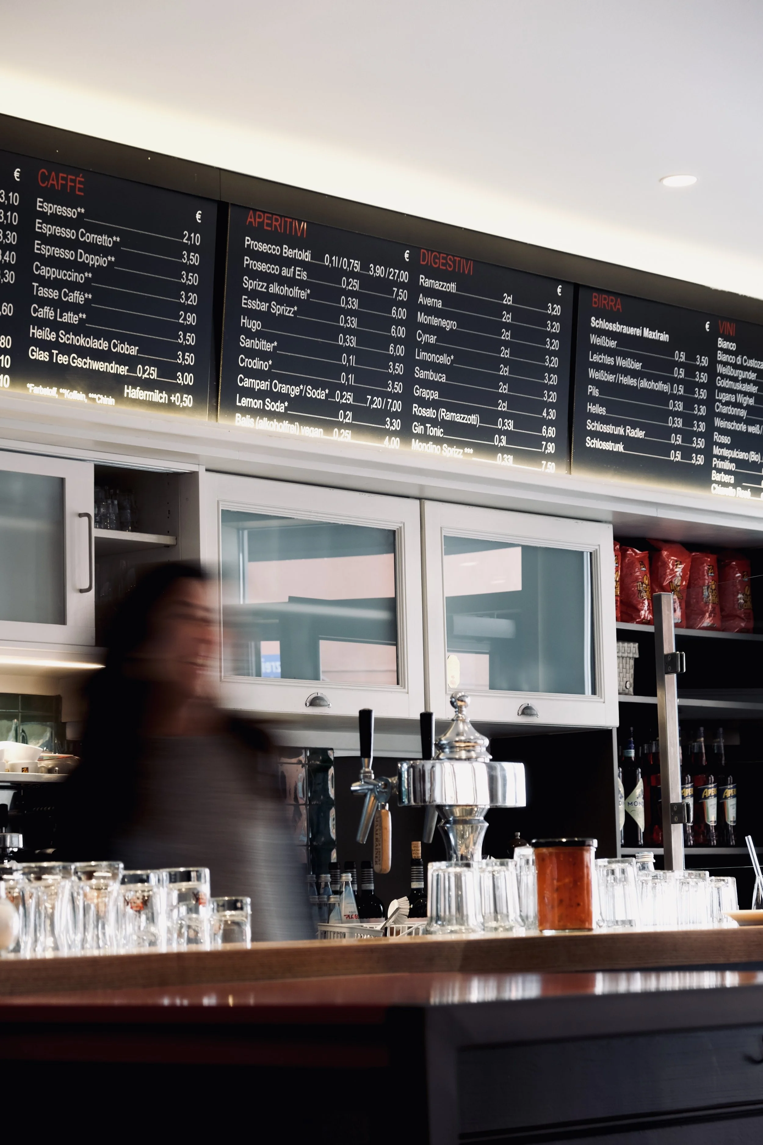 Innenaufnahme eines Cafés mit bestelltem Getränkeboard und Barista. Das Menü ist auf einer digitalen Tafel sichtbar, mit Kategorien wie Kaffee, Aperitivi, Digestivi, Bier und Wein, sowie den entsprechenden Preisen. Aufgenommen von Hilal Studios.