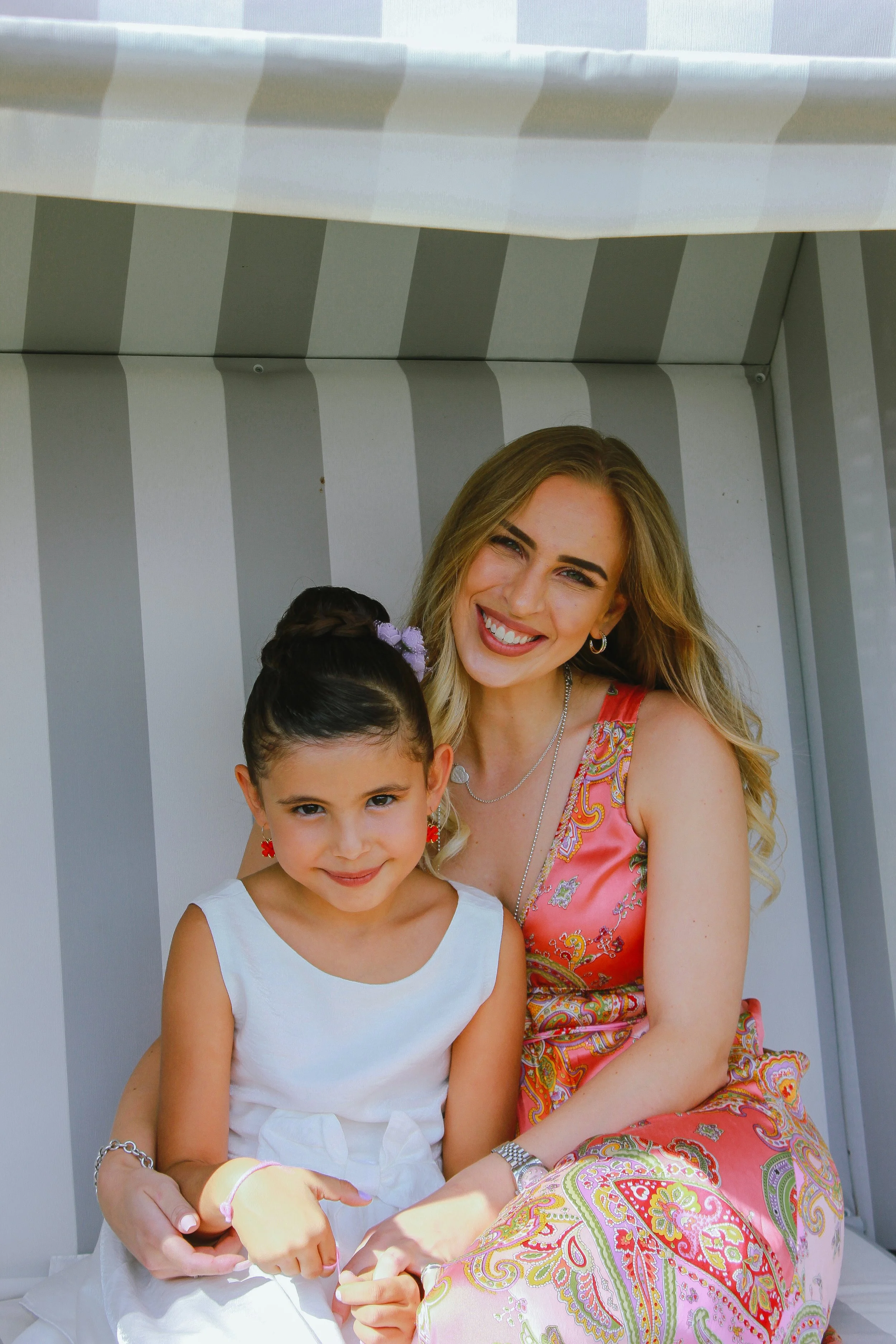 Zwei lachende Frauen, eine ältere und eine jüngere, sitzen zusammen vor einem grau-weißen gestreiften Hintergrund. Die ältere Frau trägt ein buntes, detailliertes Kleid, während die jüngere in einem weißen Kleid sitzt. Aufgenommen von Hilal Studios.