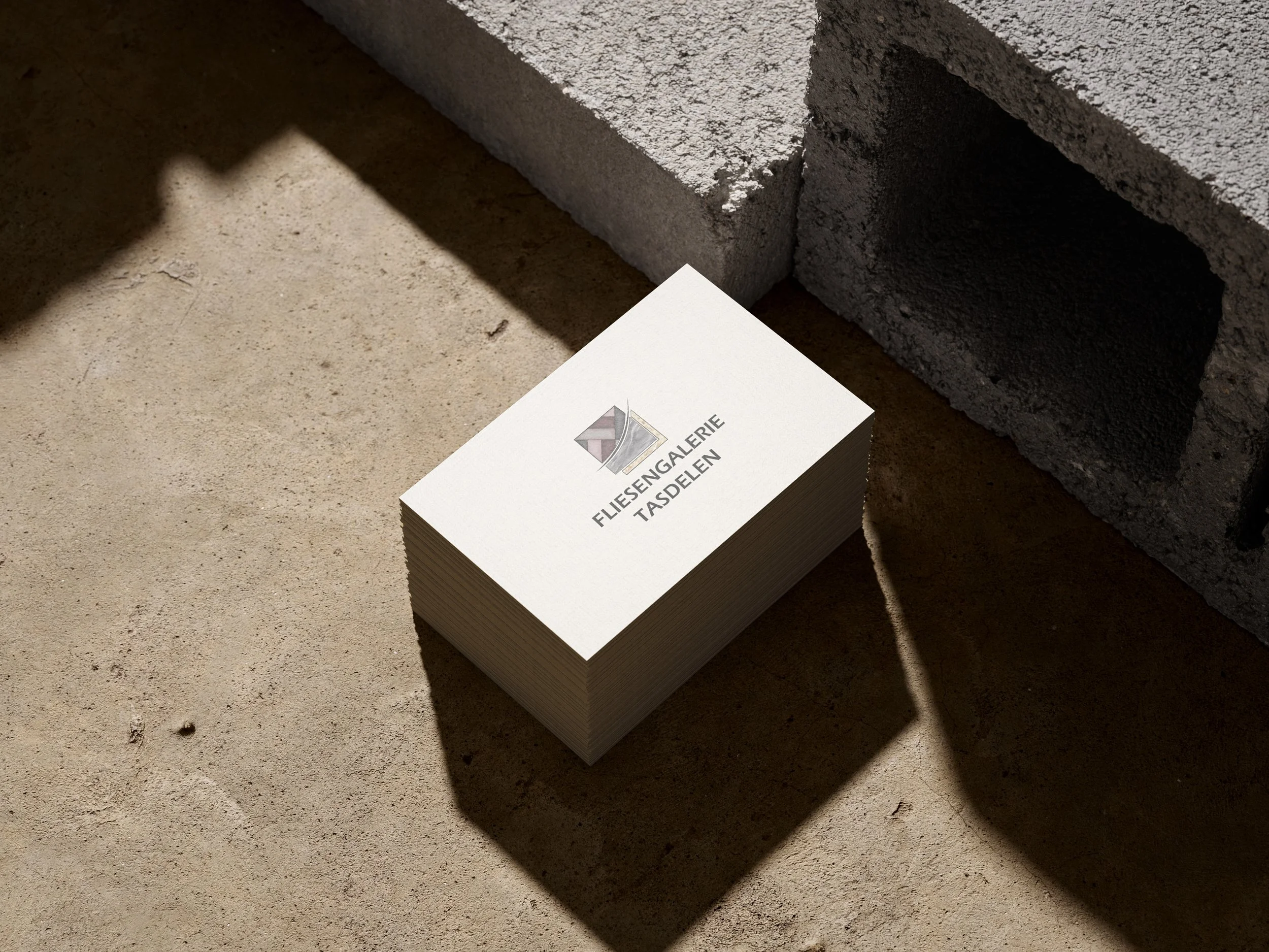 Stapel Visitenkarten mit dem Logo und Text 'Fliesengalerie Tasdelen' auf einem sandfarbenen Boden neben einem Ziegelmauerstück. Erstellt von Hilal Studios.