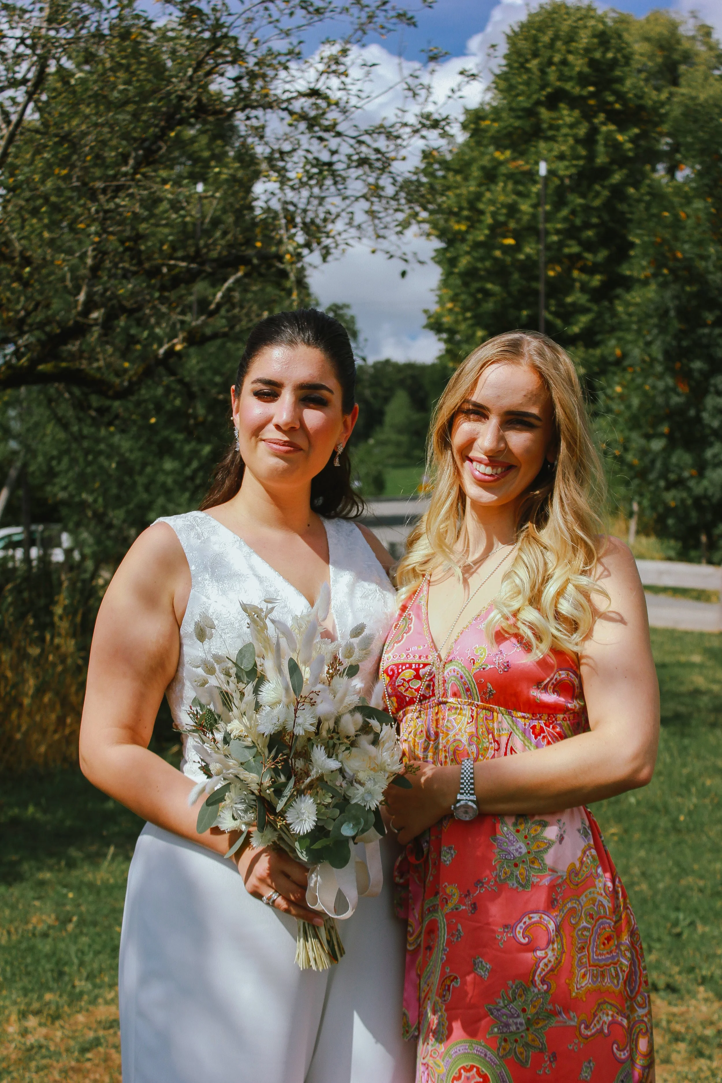 Zwei Frauen im Freien, eine in einem weißen Kleid mit Brautstrauß, die andere in einem bunten Kleid, beide lächeln bei sonnigem Wetter. Aufgenommen von Hilal Studios.