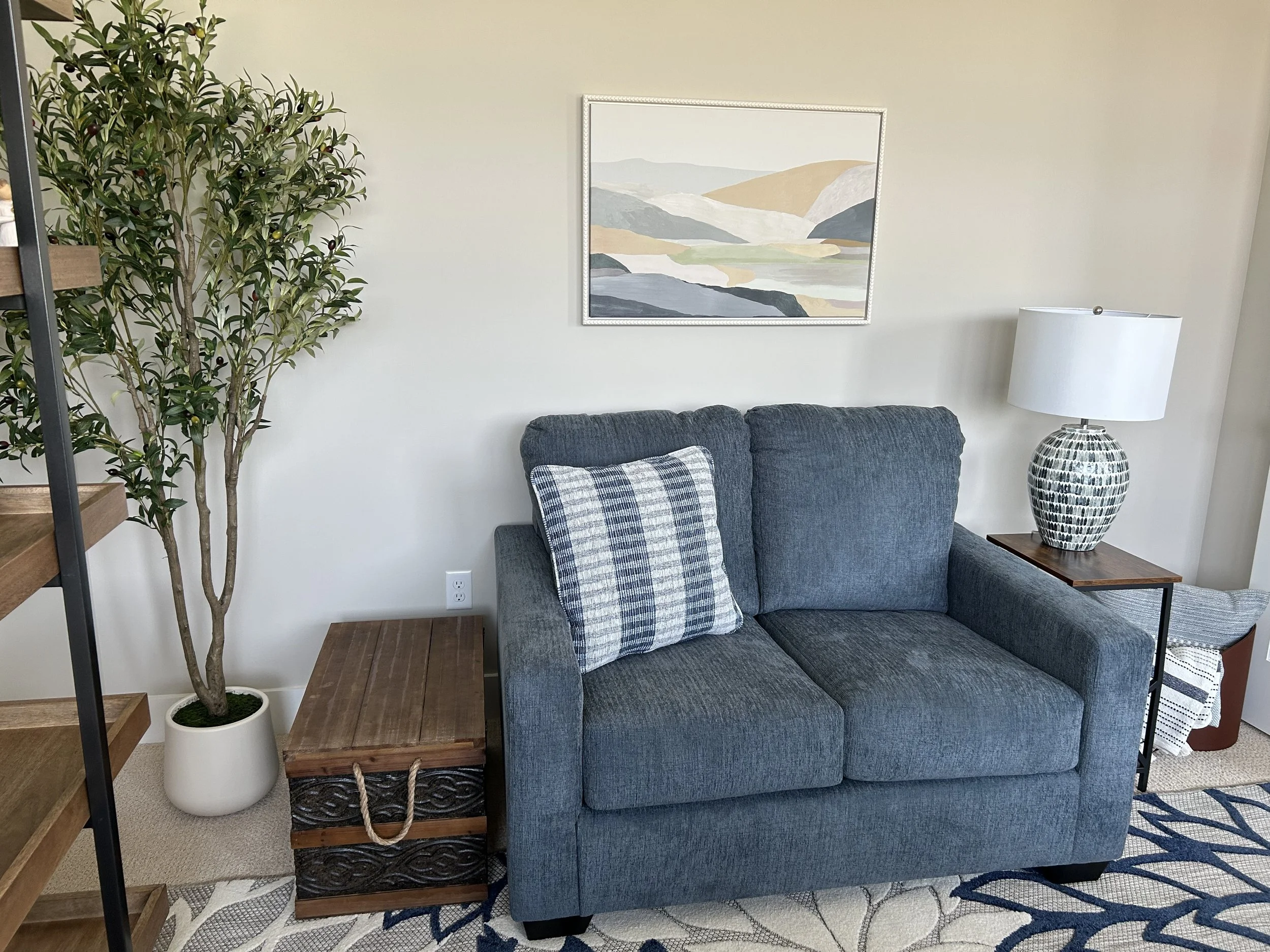blue loveseat, faux olive tree, wooden storage trunk, glass mosaic table lamp, abstract mountain wall art