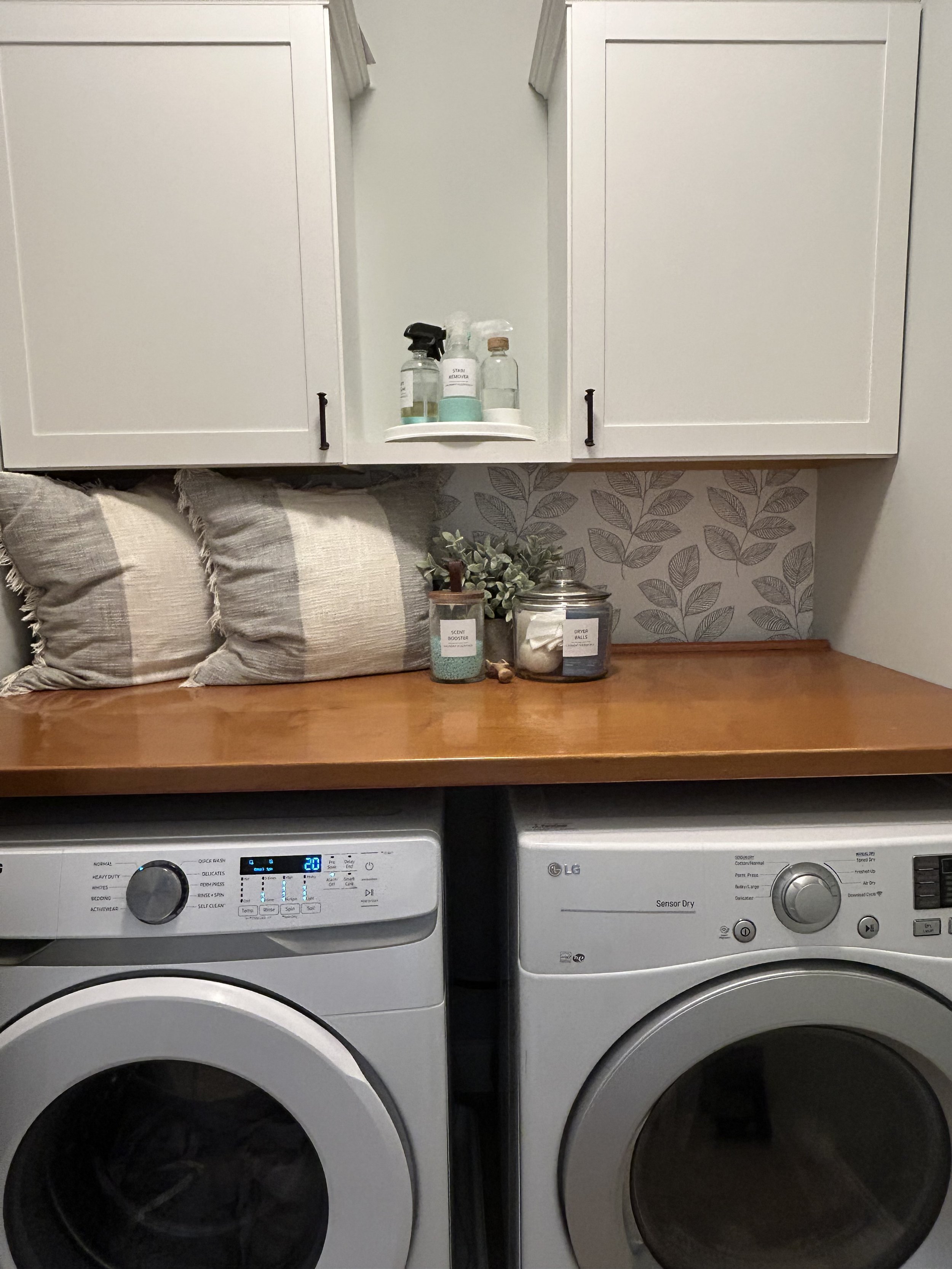 The wallpaper and throw pillows add some personality and coziness to this clean, fresh laundry room space. We've decanted all of the cleaning products into coordinated labeled containers. We kept items we use frequently easily accessible, and backsto