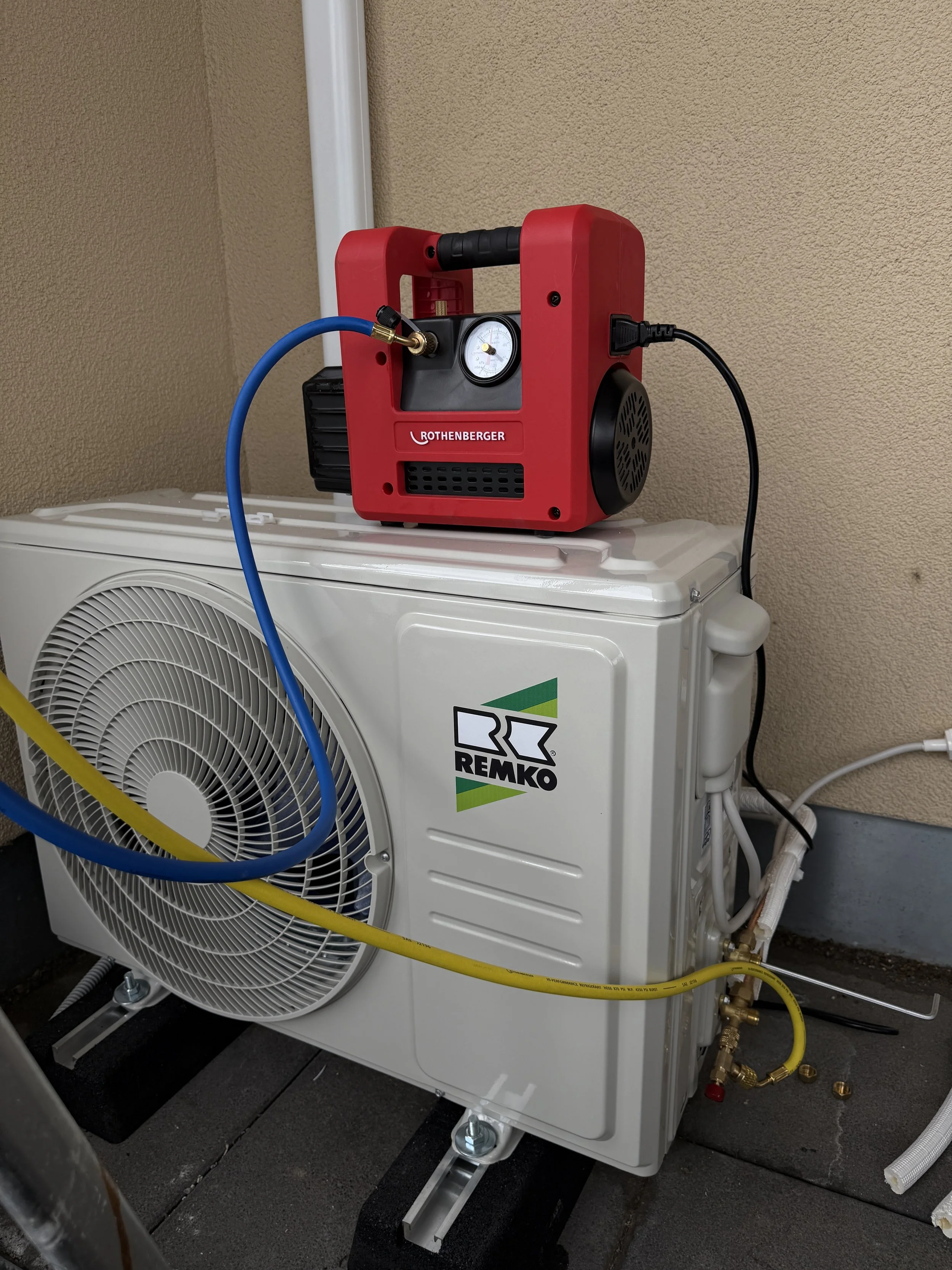 Ein rotes Gerät, wahrscheinlich eine Pumpe oder ein Kompressor, steht auf einer weißen Außen-Klimaanlagen-Einheit mit der Marke REMKO. Das Gerät ist an die Einheit angeschlossen und befindet sich in einem technischen oder Wartungsbereich.