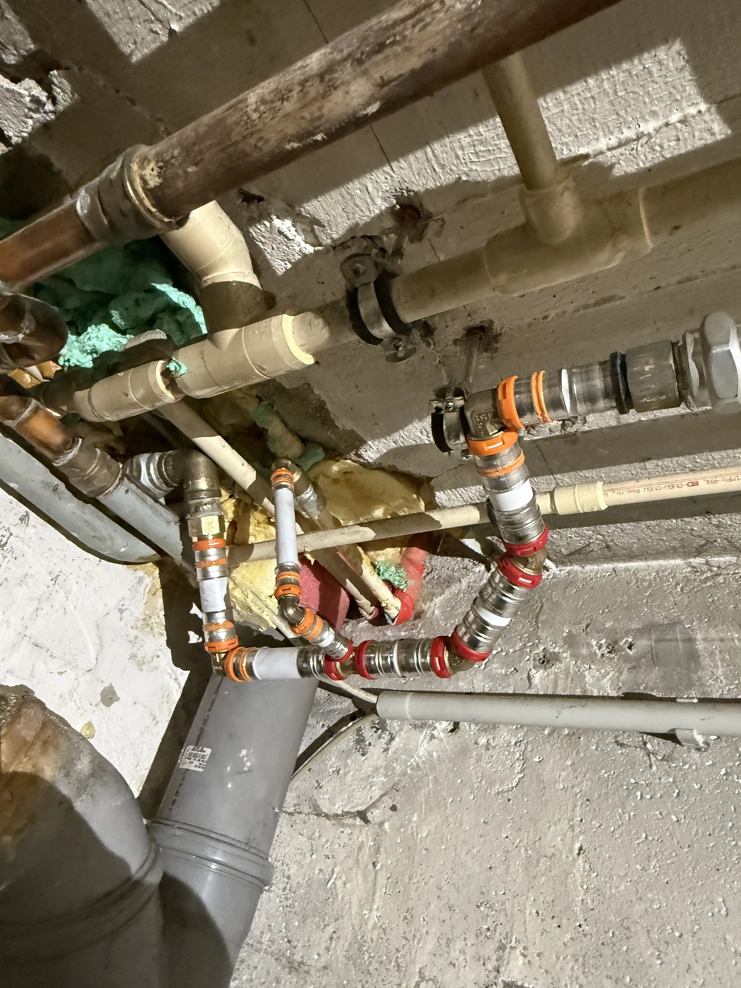 Unterschiedliche Wasserleitungen in einem Keller, darunter Kupferrohre, Kunststoffrohre und ein Fallrohr mit Verbindungsteilen.