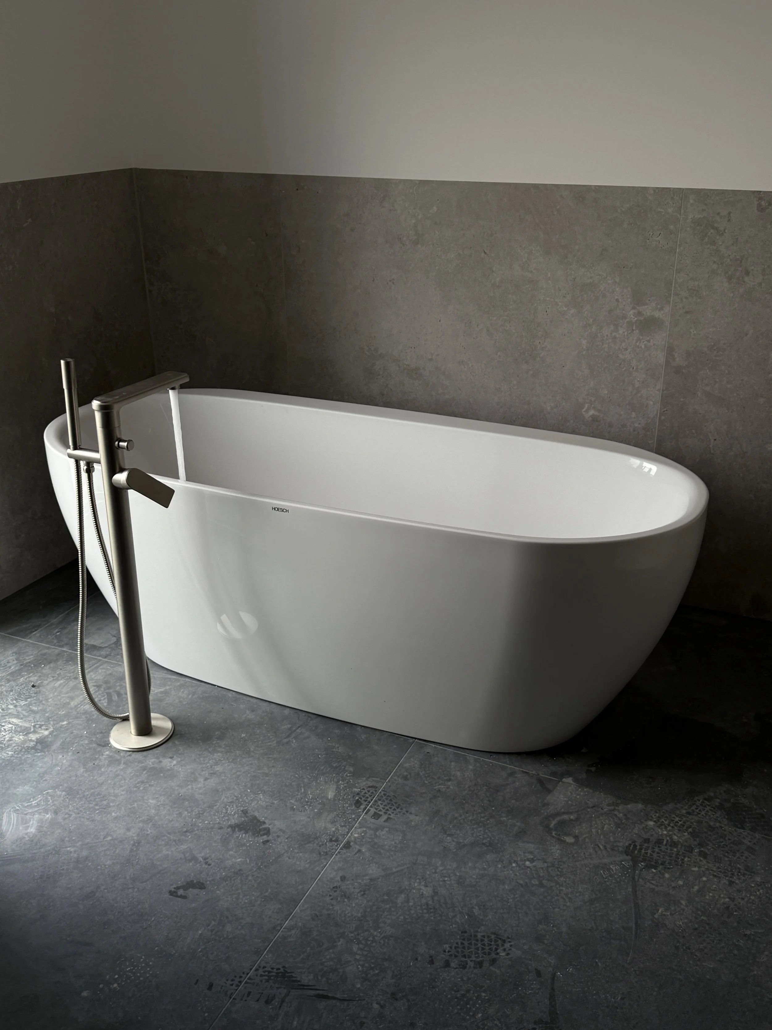 Moderne weiße freistehende Badewanne in einem Badezimmer mit dunklen Fliesen und grauem Wandstück