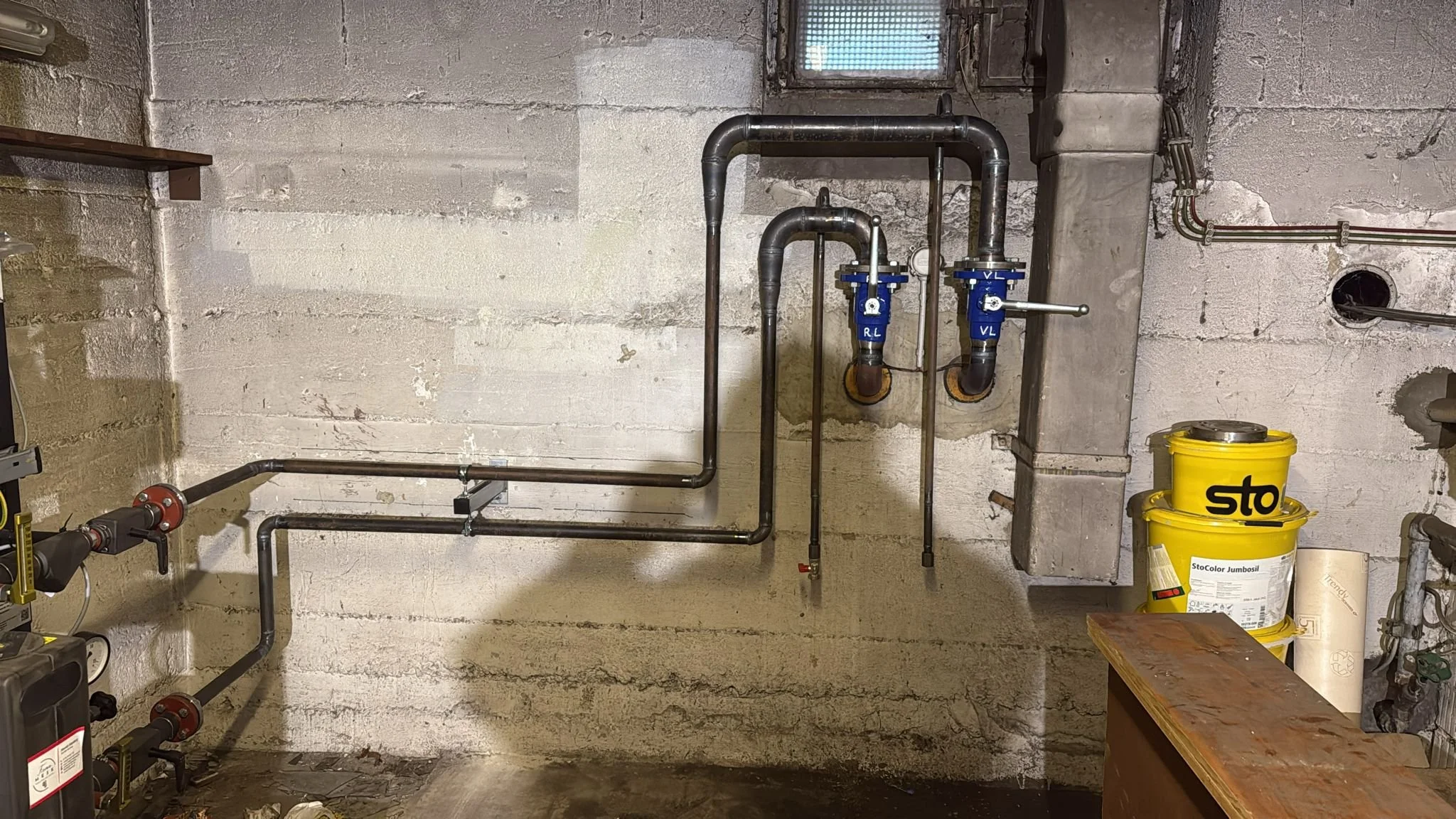 In einem Keller oder Technikraum sind schwarze Rohrleitungen mit Wasserhähnen, Absperrventilen und Messgeräte an einer Wand montiert. Auf der rechten Seite befinden sich zwei gelbe Farbdosen, ein Holzarbeitsplatte und ein Rohr.