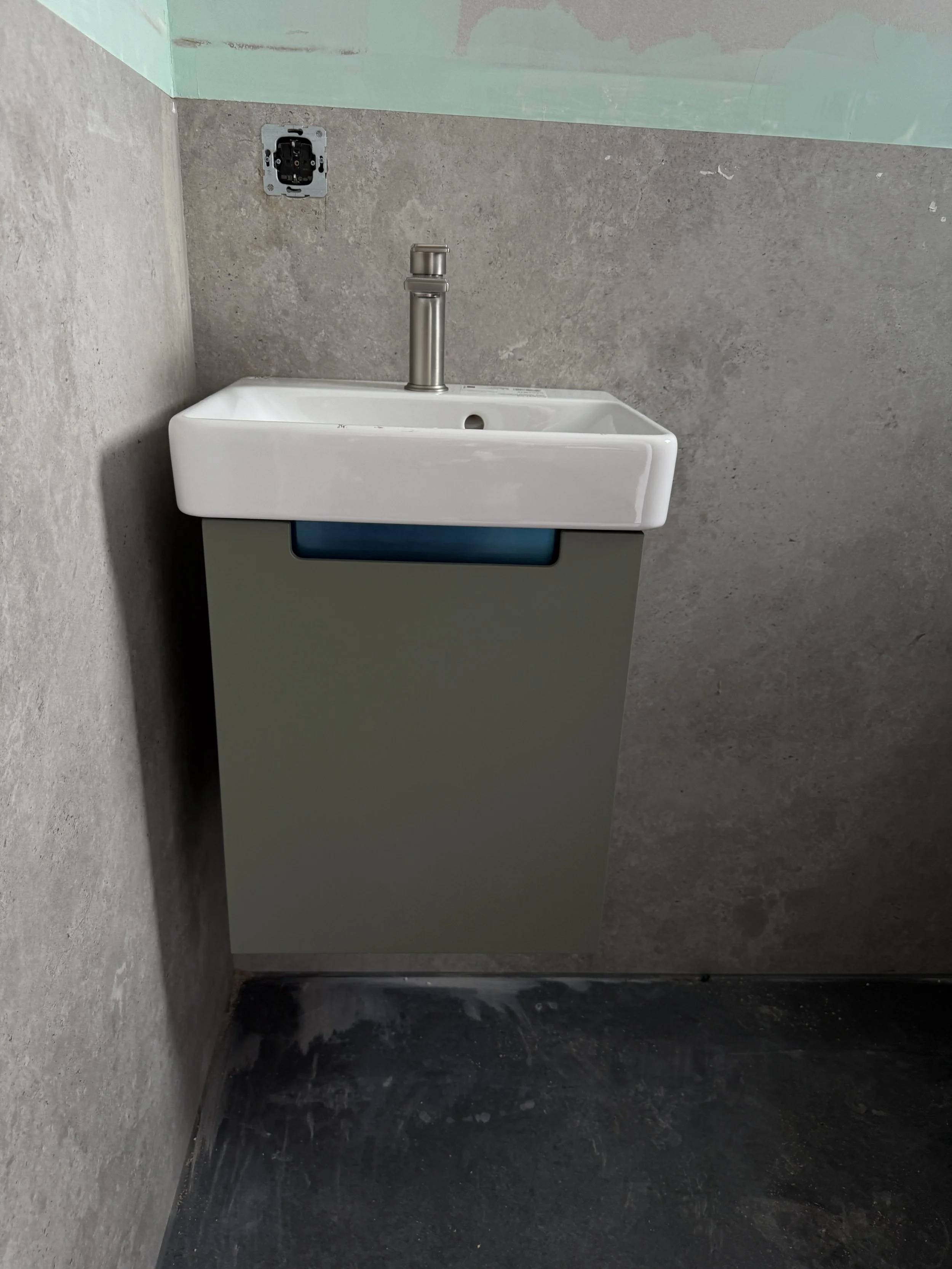 Ein weißes Handwaschbecken auf einem grauen Unterschrank, installiert in einer Ecke einer dünnen Wand aus grauem Beton, ohne Wasserhahn, nur mit einem Anschlussrohr.