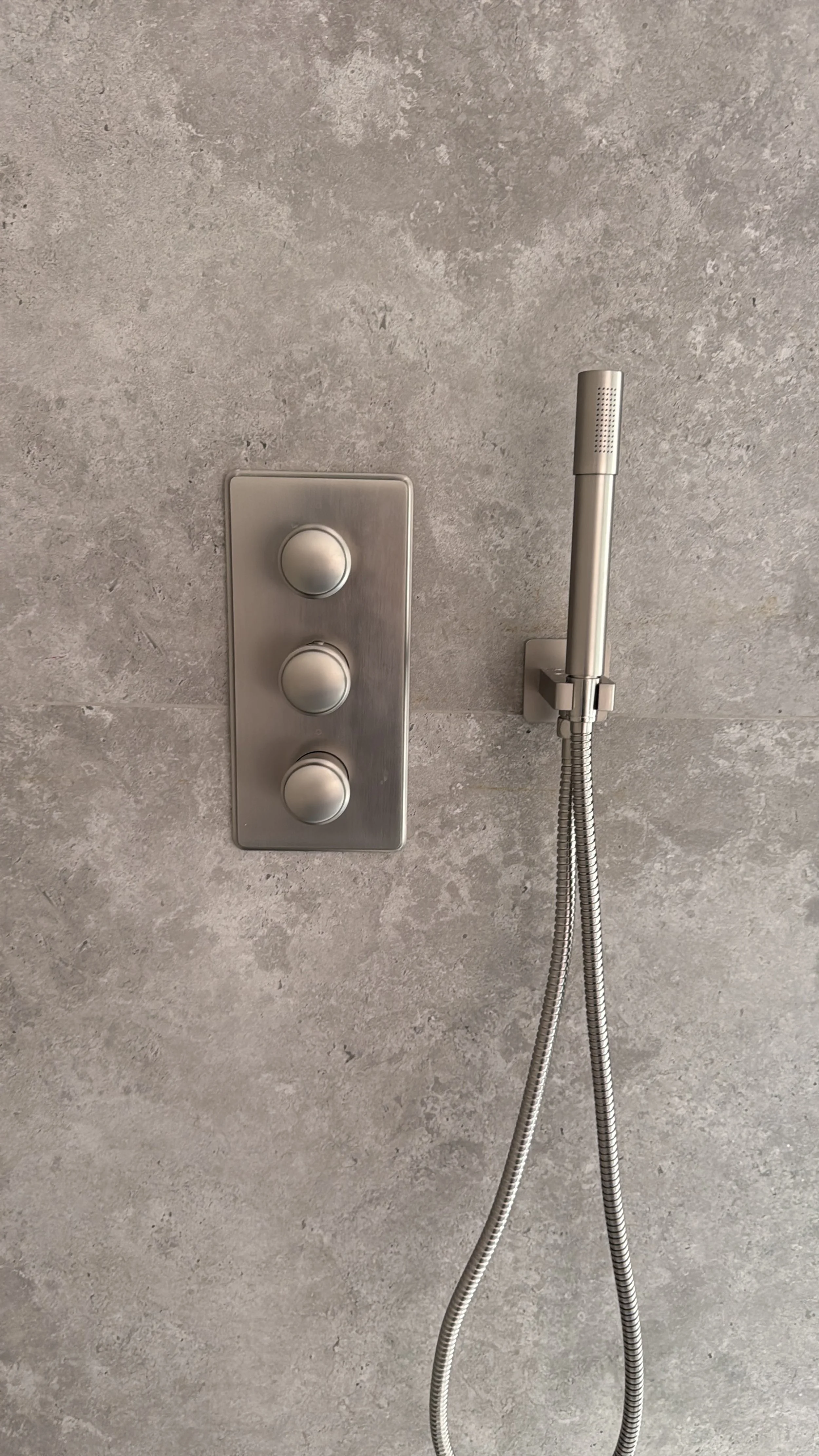 Ein Duschbereich mit grauem Wandfliesen, einem Duschknopf mit drei Druckknöpfen und einem Handbrausekopf mit flexiblem Schlauch.