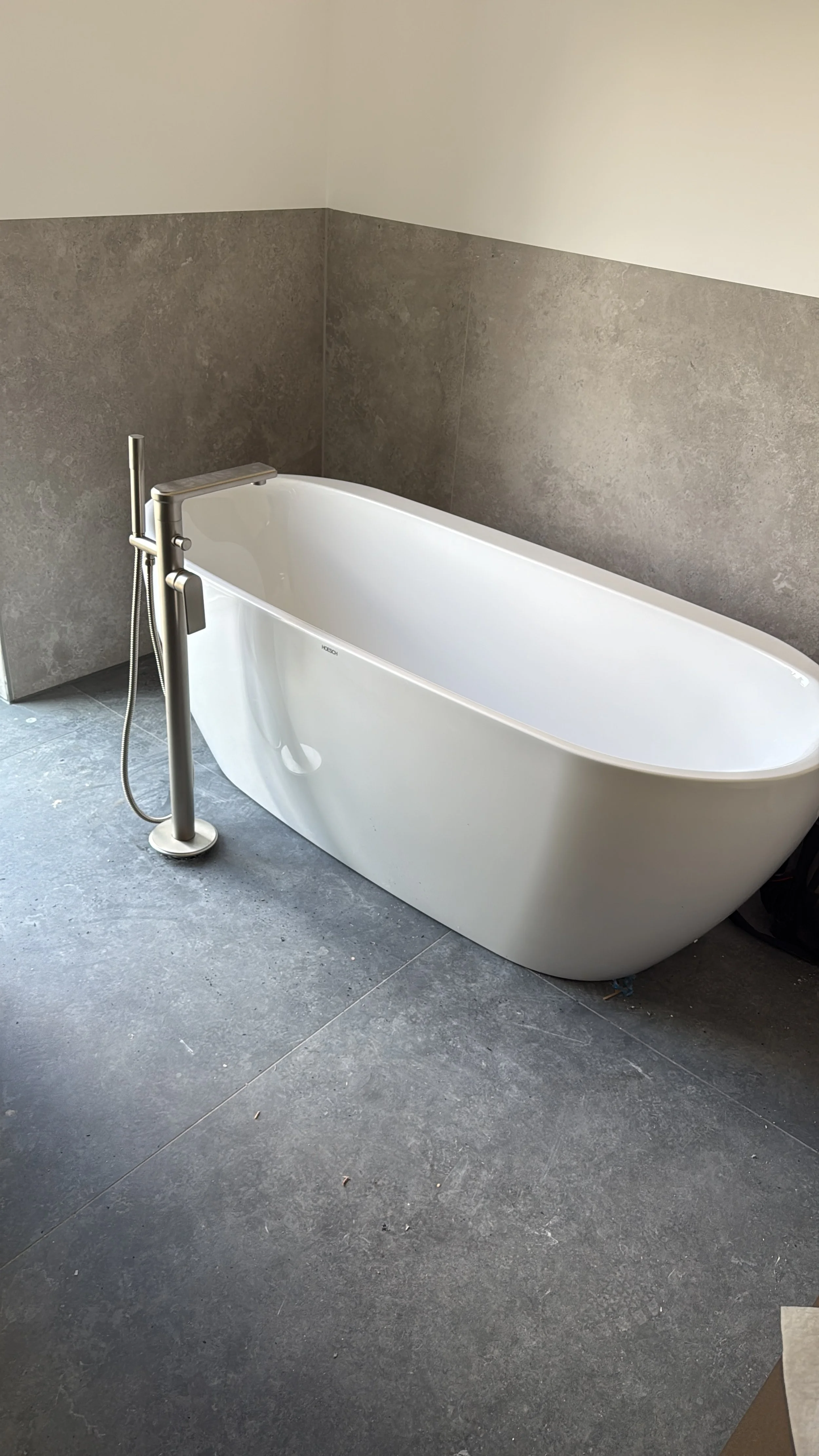 Weiße Badewanne mit moderner Armatur in einem Badezimmer mit grauen Fliesenboden und breiten hellgrauen Wänden.