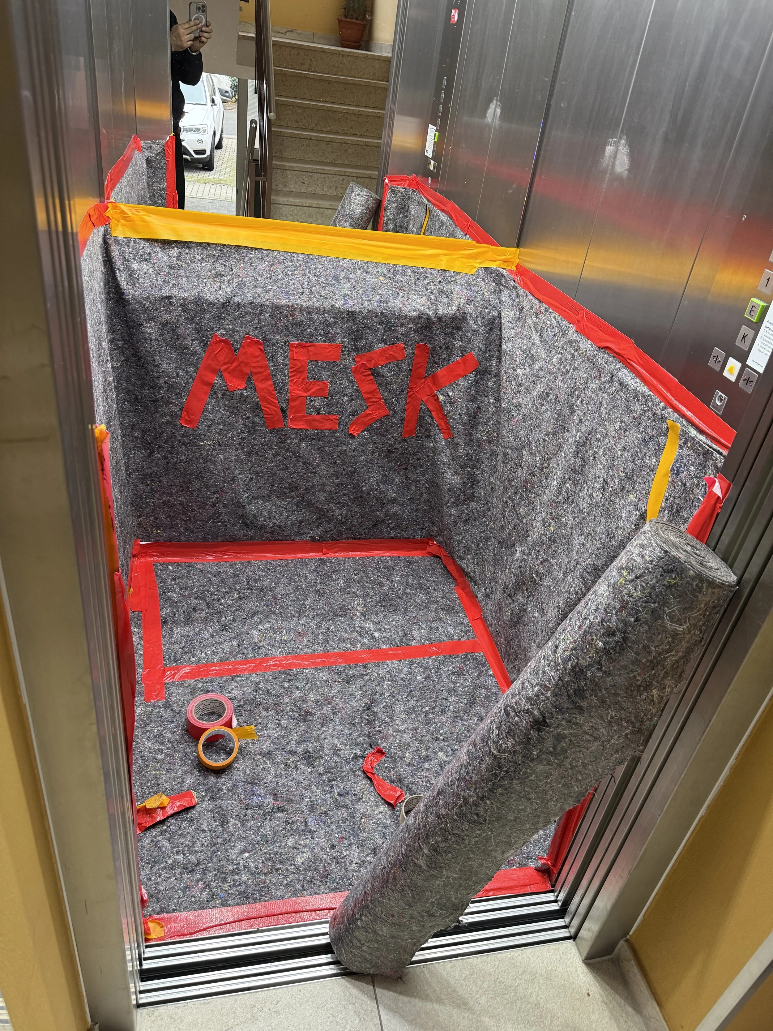 Ein aufgeräumter Aufzug mit Baustoffen, darunter Isoliermatten, Klebebänder und rotes Malerband, sowie eine aufgemalte Aufschrift 'MEKA' an der Wand.