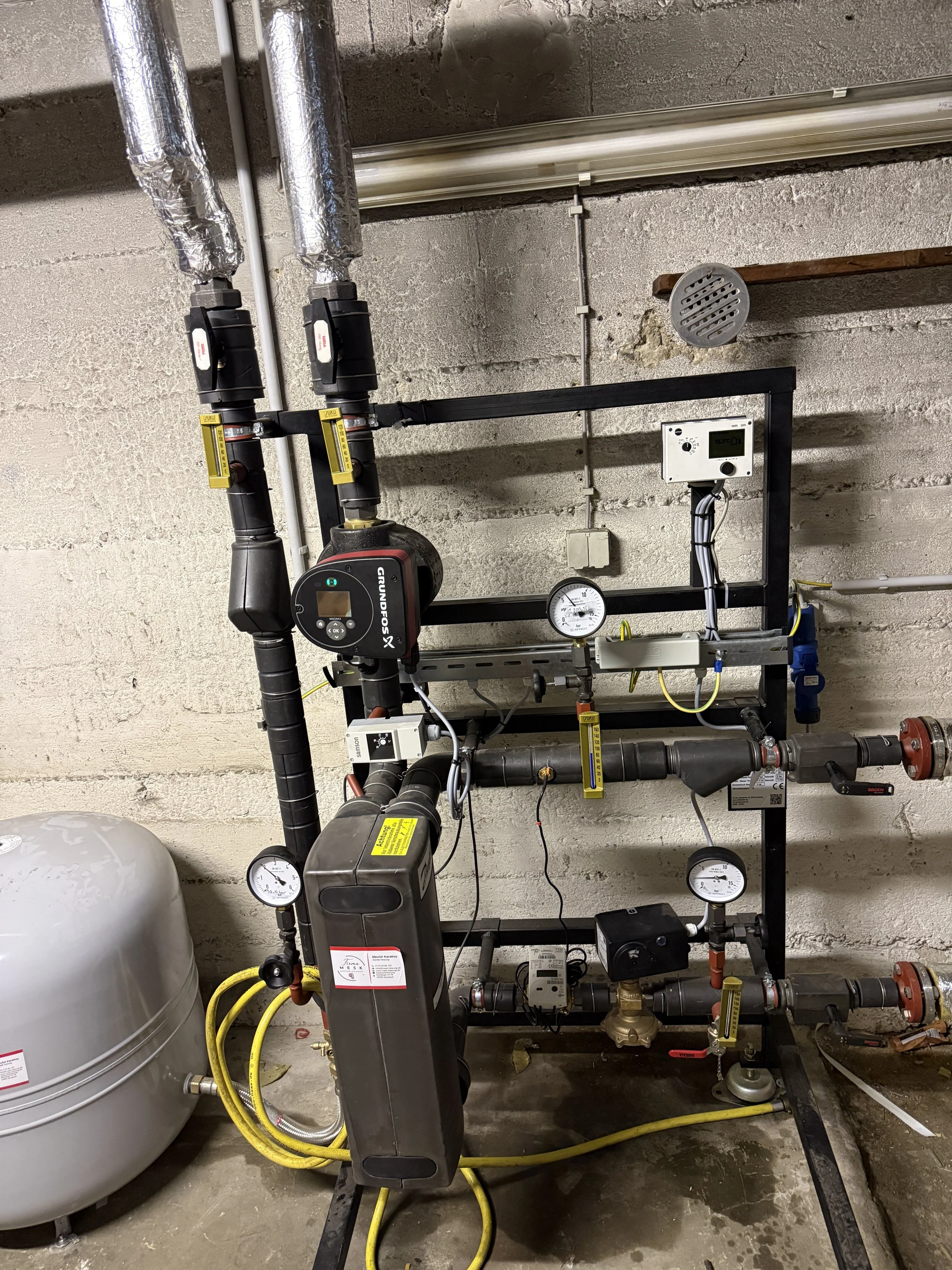 Technische Anlage mit Ventilen, Pumpen und Messgeräten in einem Kellerraum.