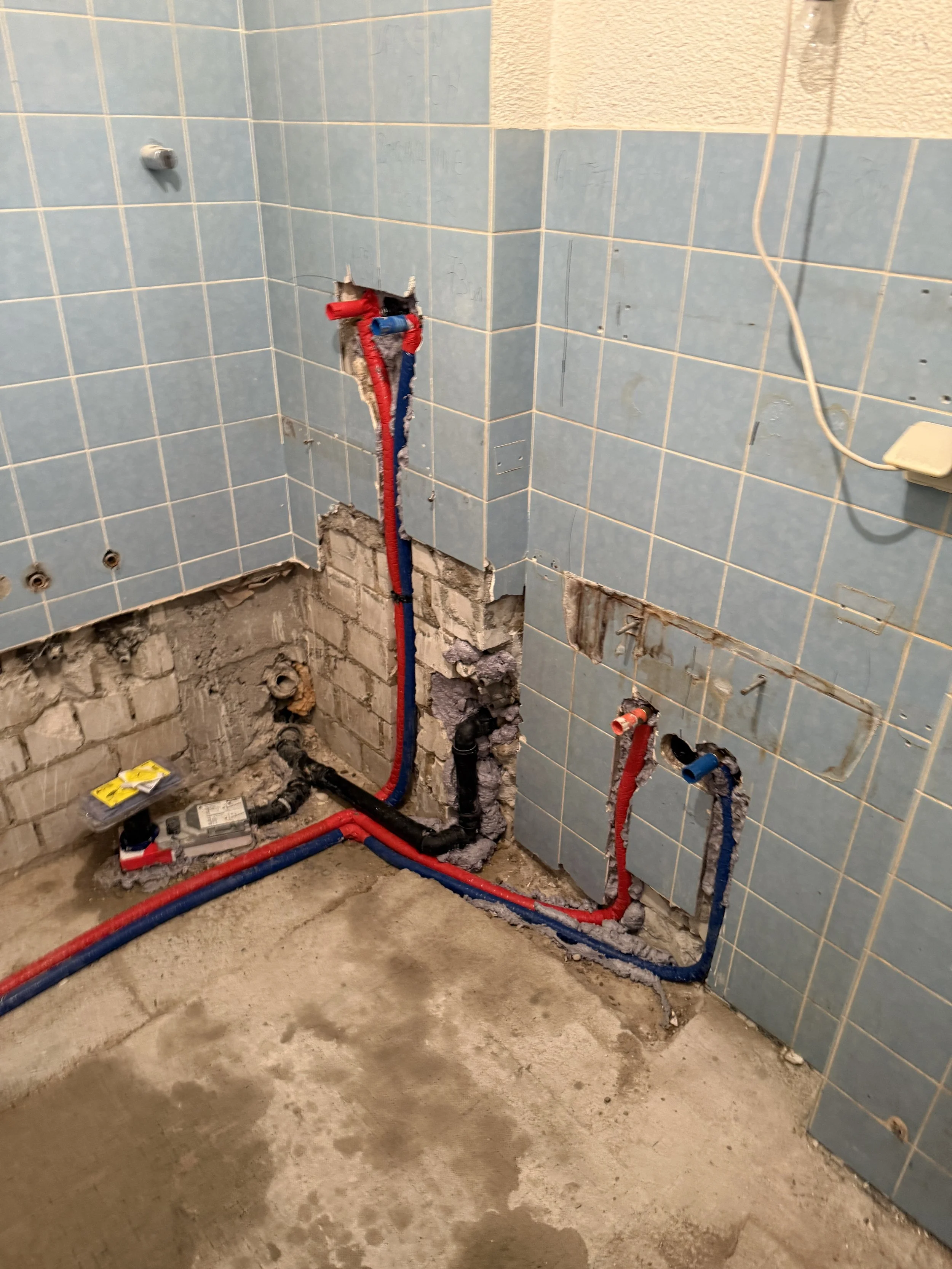 Badezimmer während der Renovierung mit sichtbaren Wasserleitungen in Rot und Blau, die für Warm- und Kaltwasser stehen. Die Wand ist teilweise offen, um die Rohre zu installieren, und die Fliesen sind teilweise entfernt.