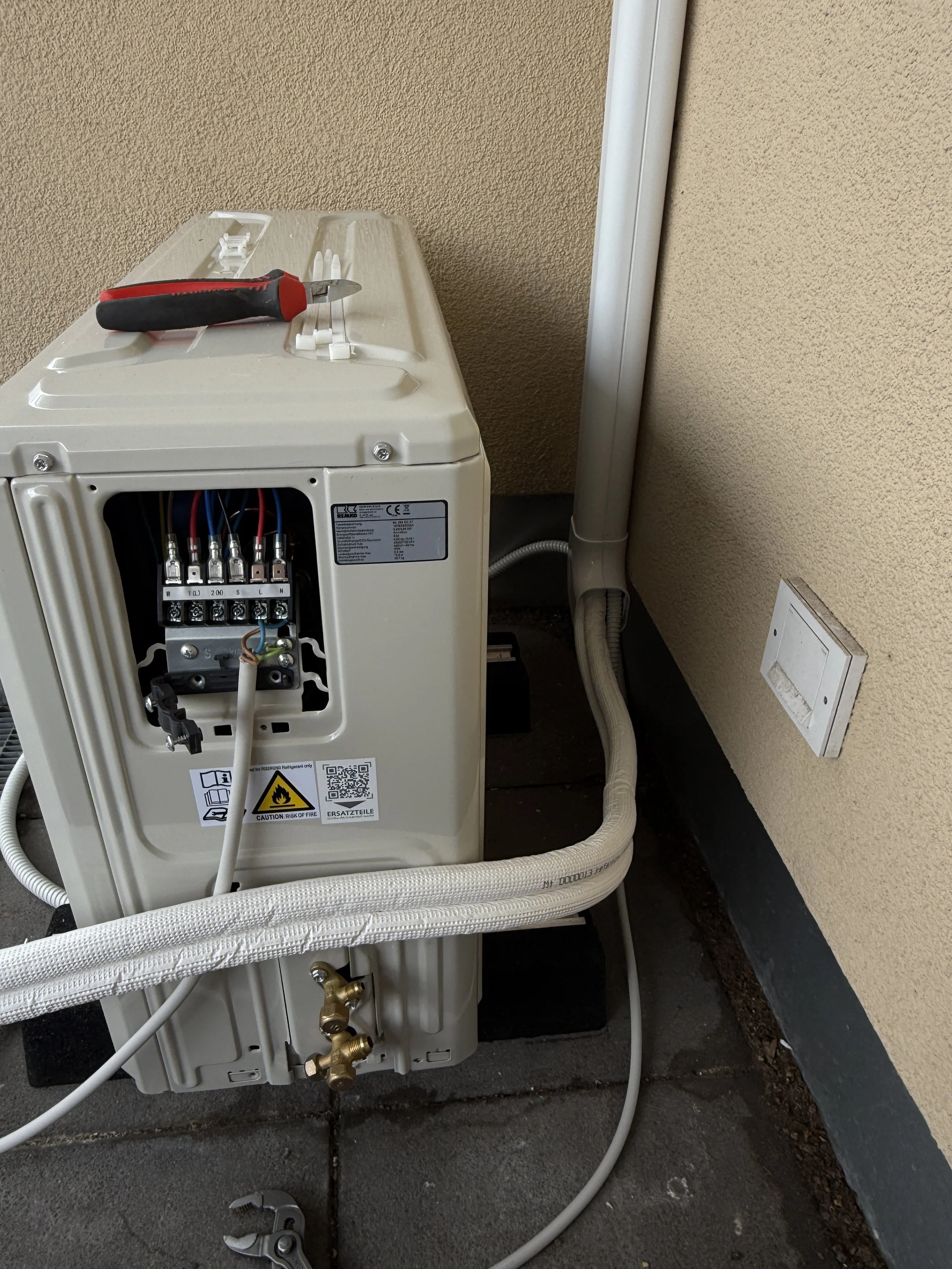 Ein offener Schaltschrank mit einem Schraubendreher auf der Oberseite, mehrere Kabel und Drähte sowie Verbindungsklemmen, sich neben einer Wand mit Steckdose befindend.