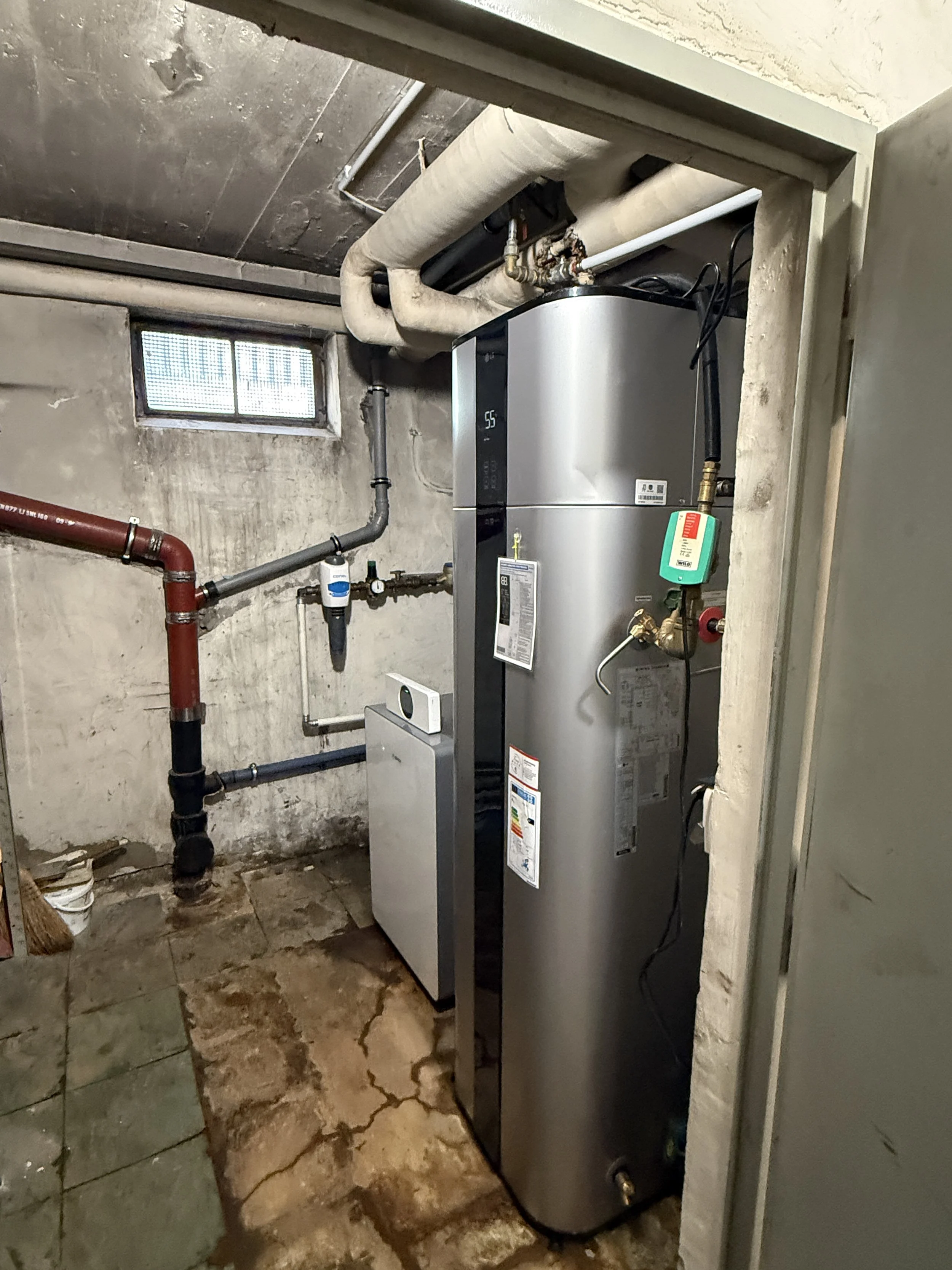 Ein Heizungsraum mit einer modernen, panelförmigen Heizungseinheit, Rohrleitungen und einem kleinen Fenster.