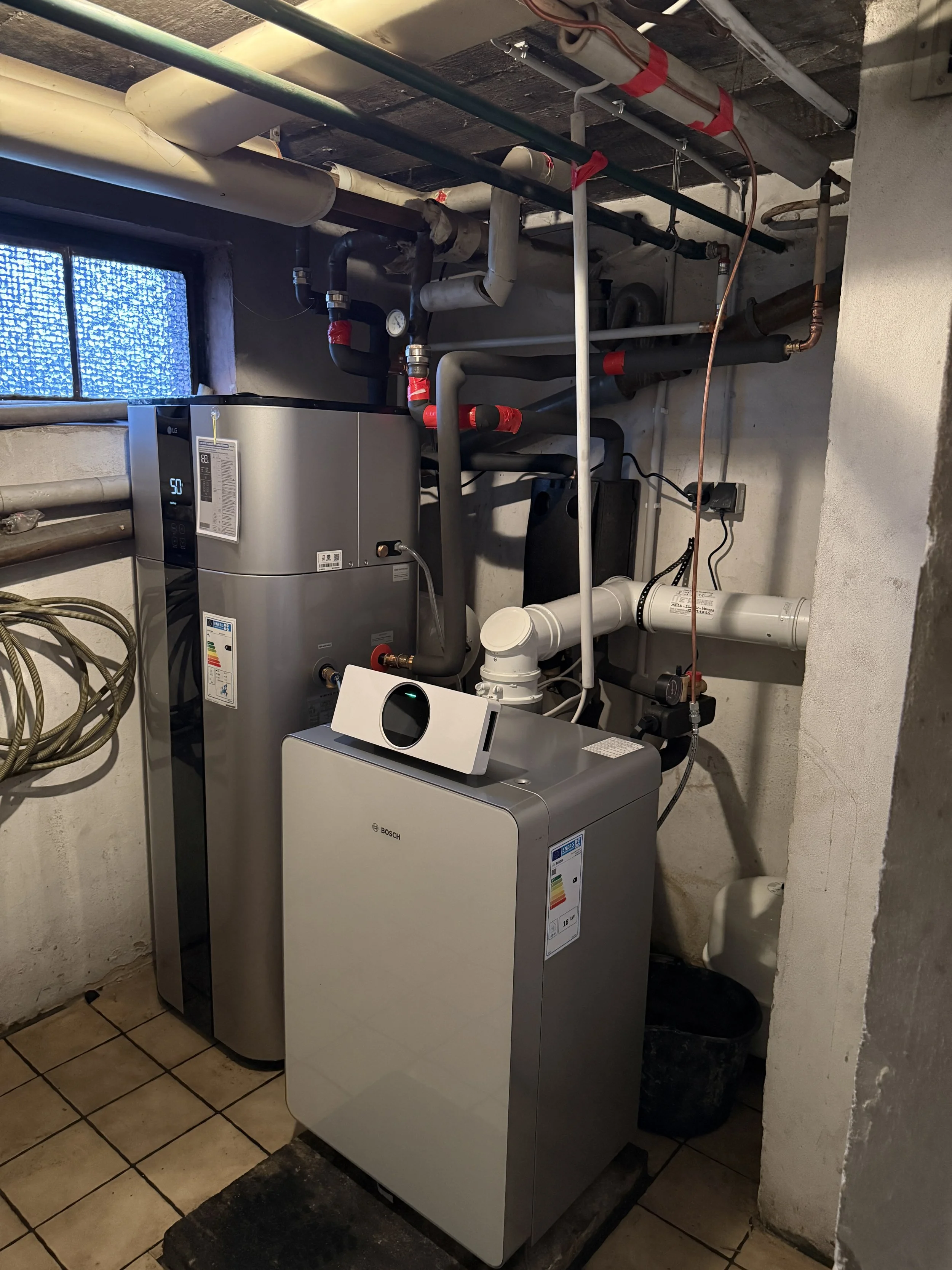 Ein Hauswirtschaftsraum mit einer modernen Wärmepumpe und einer Heizungsanlage, umgeben von Rohrleitungen und Wasserleitungen, auf Fliesenboden mit kleiner Fenster im Hintergrund.