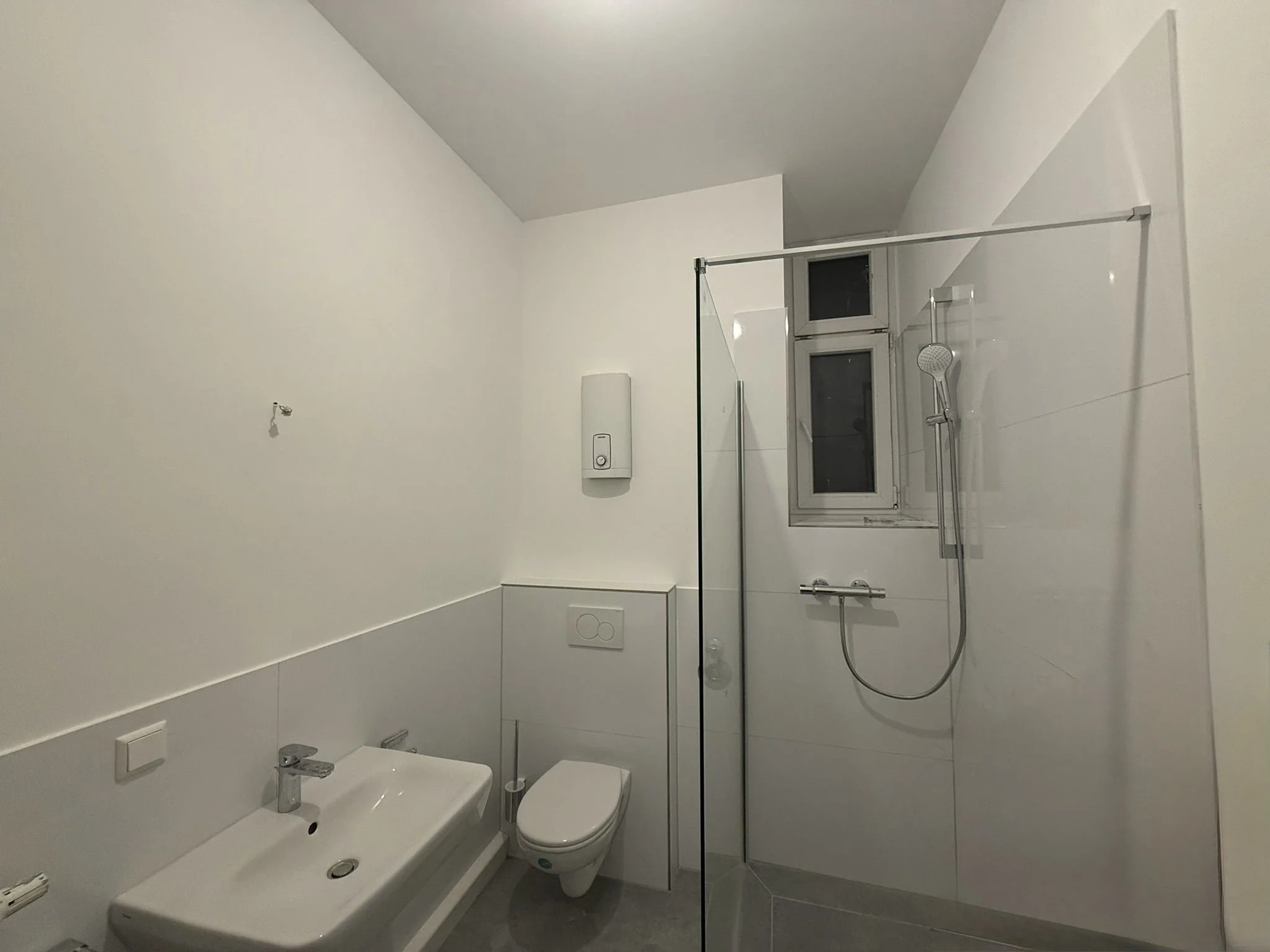 Ein modernes Badezimmer mit weißem WC, Waschbecken, ebenerdiger Dusche mit Glastrennwand, Fenster und weißen Fliesen.