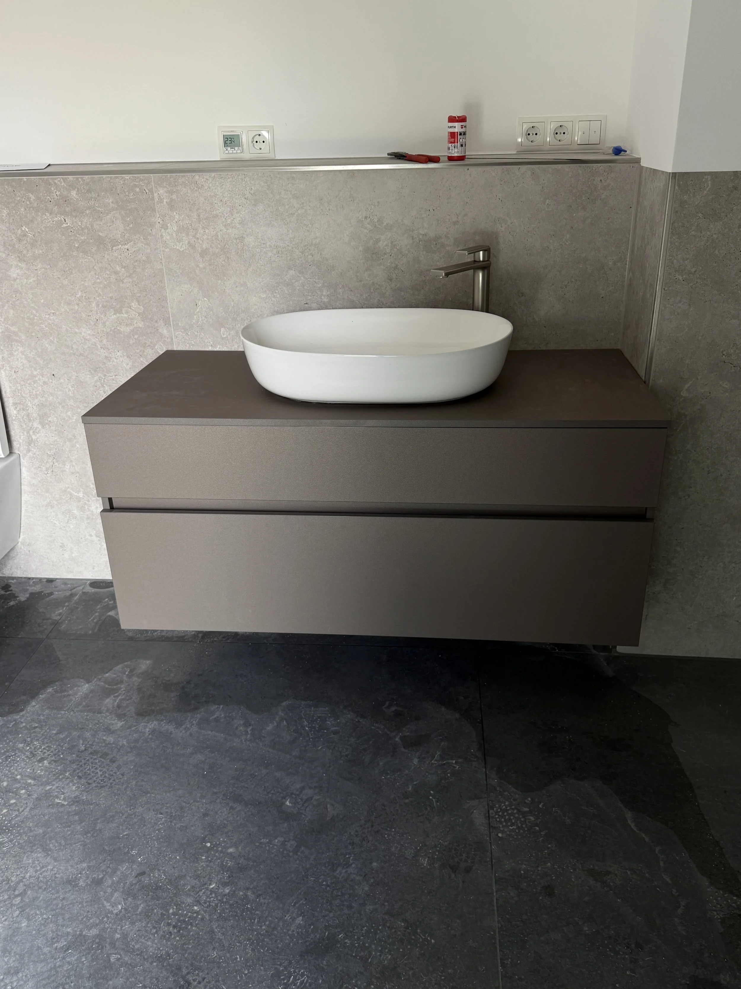 Ein Waschbecken auf einem grauen Waschtisch in einem Badezimmer, Wandfliesen im Hintergrund, mehrere Stromanschlüsse und Werkzeuge auf der Wandleiste.