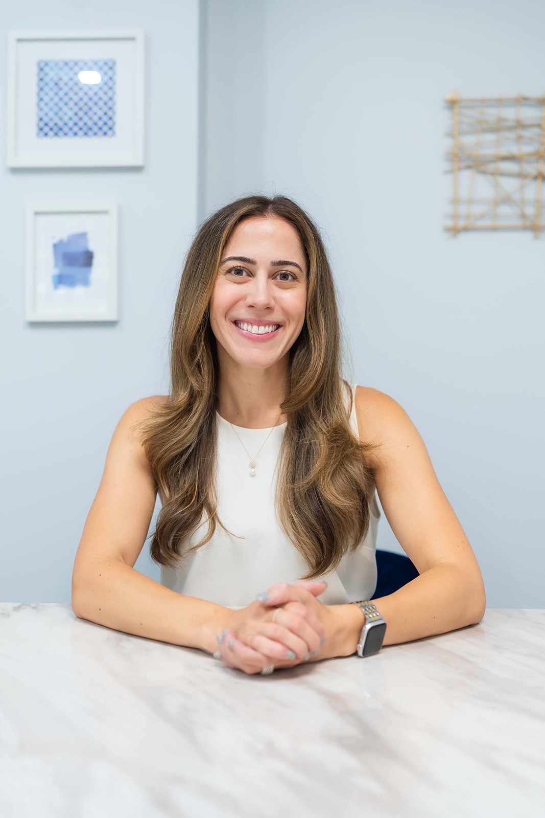 Woman smiling and seated at a marble table with art on the wall in the background.