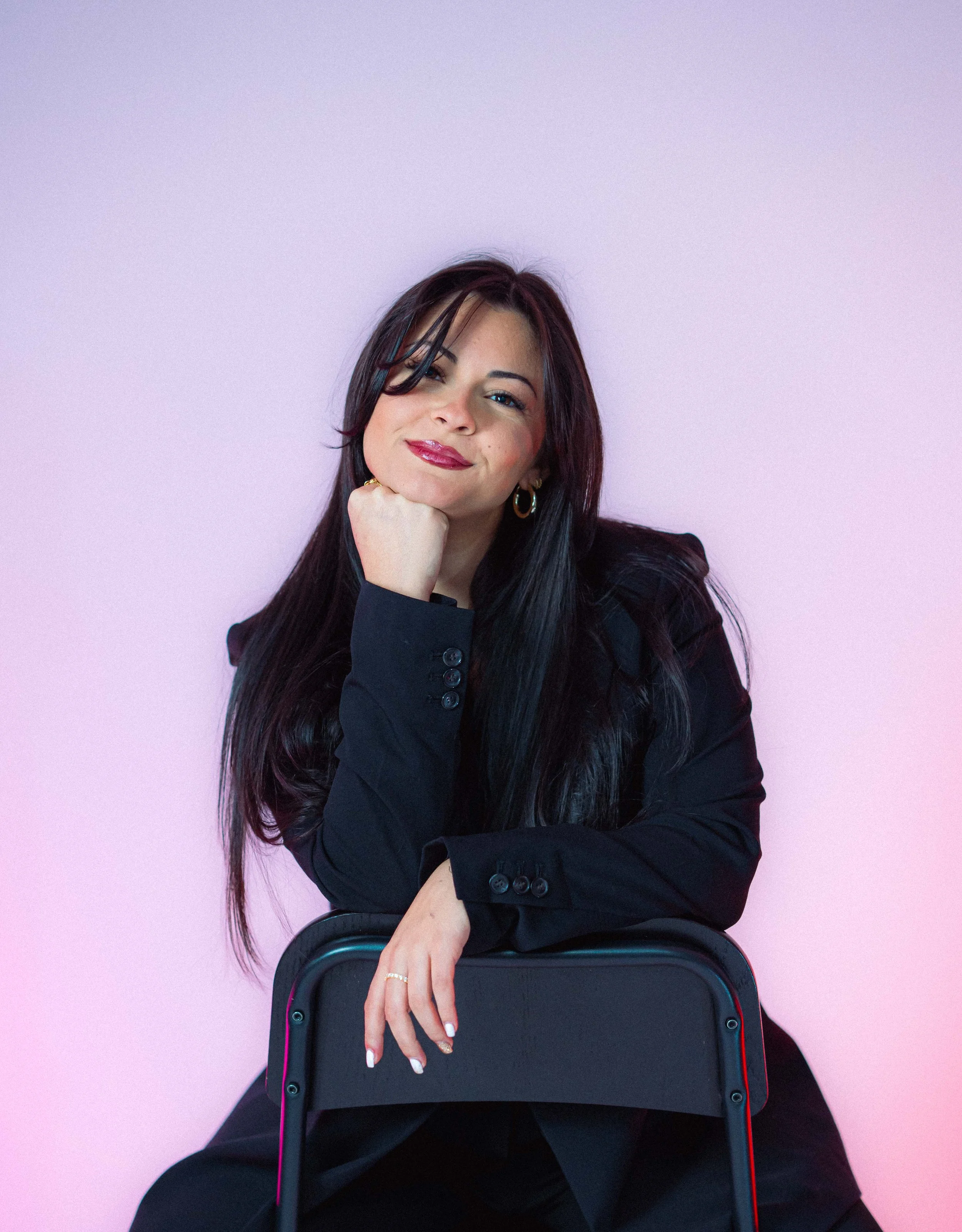 Woman in black blazer sitting on chair against a light purple background, smiling.
