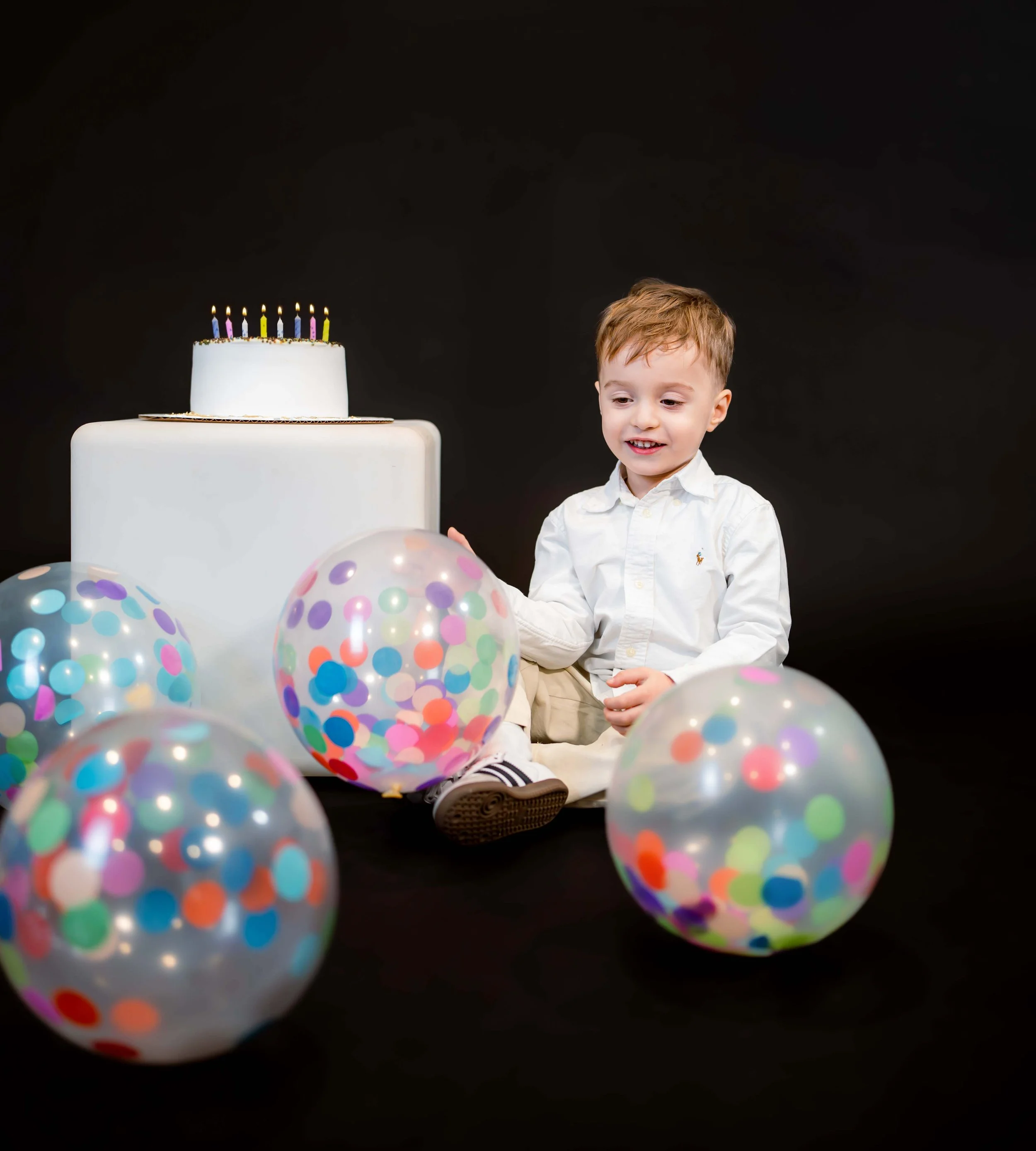 A child wearing a white shirt sits on the floor with colorful polka dot balloons around, next to a white cake with candles on a pedestal. The background is black.
