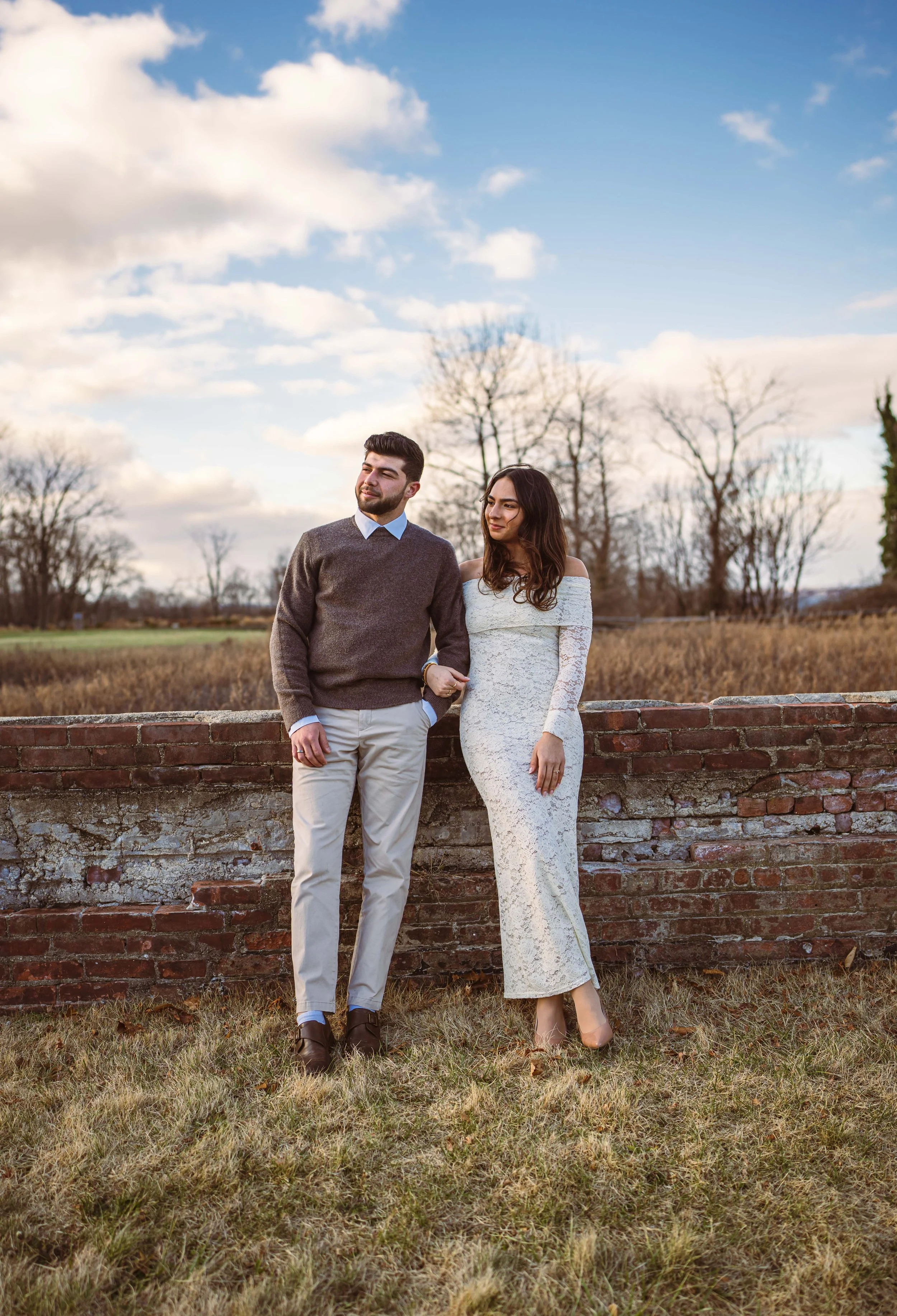 A couple standing outdoors by a brick wall, holding hands, with a grassy field and bare trees in the background. The man is wearing a brown sweater and khaki pants, and the woman is in a white lace dress.