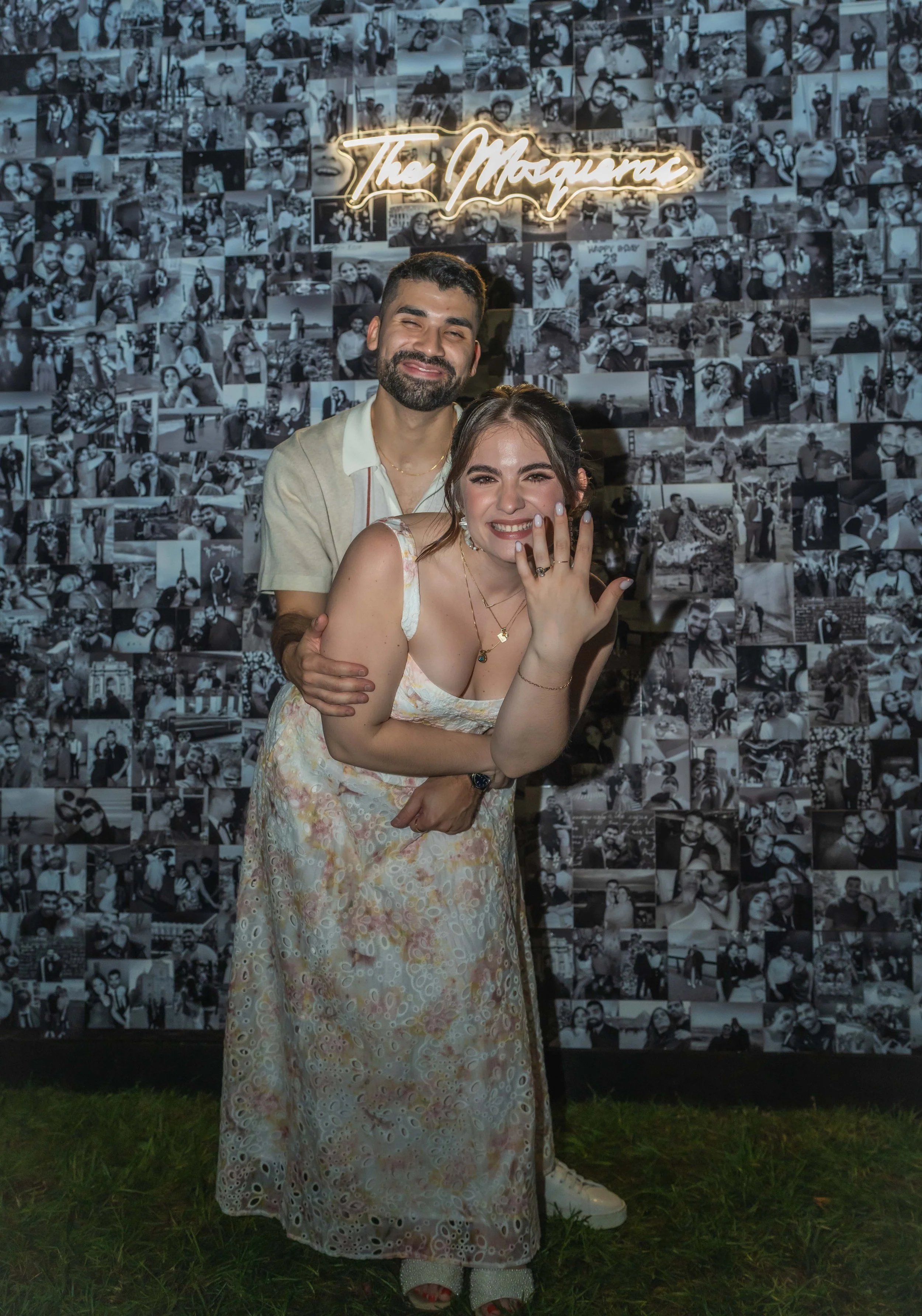 Smiling couple posing in front of a collage of black and white photos, with the woman showing off a ring. Neon sign with 'The Mosqueras' above them.