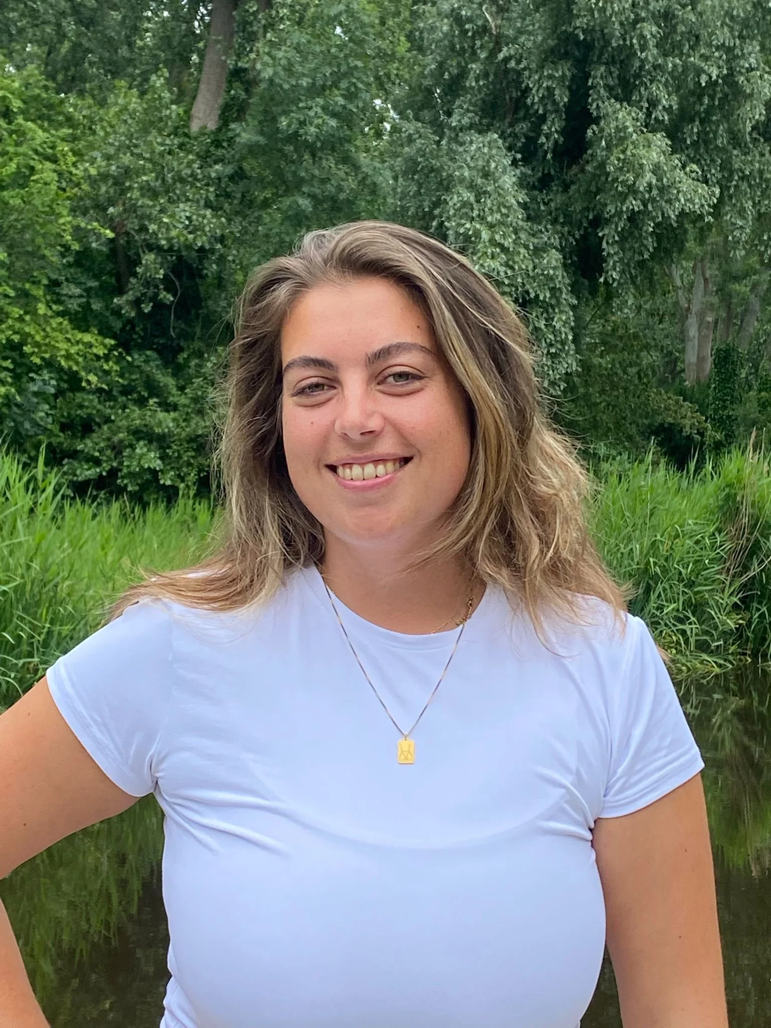 Laura, glimlachend, buiten in een groene natuurlijke omgeving met bomen en water.