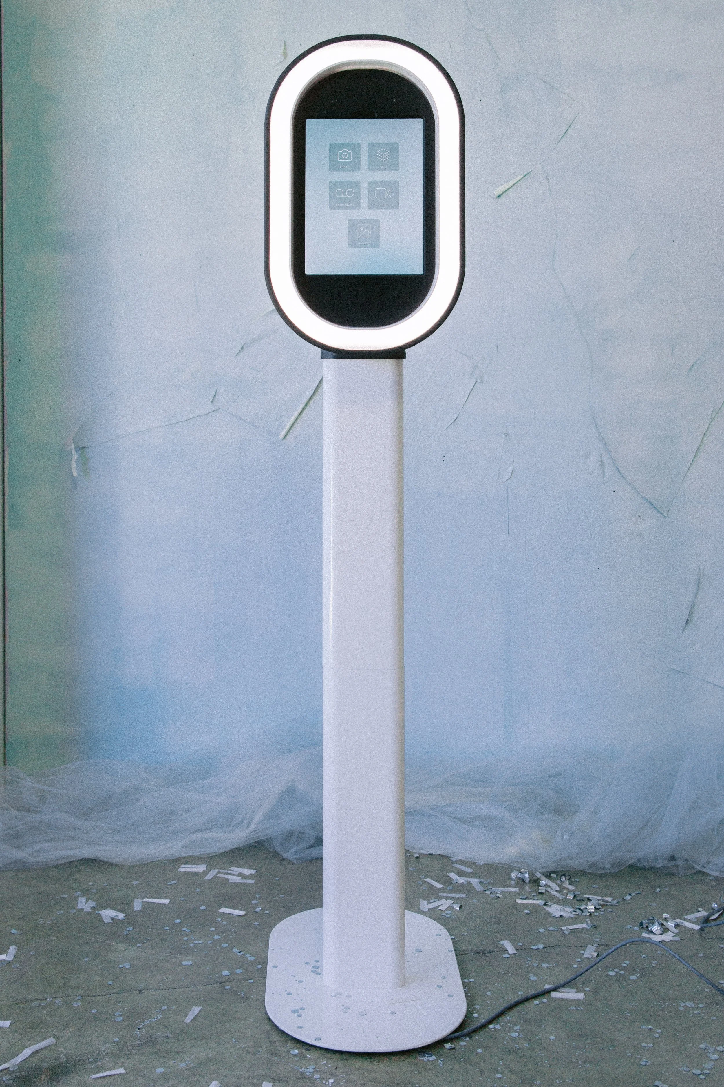 A sleek, modern touchscreen kiosk with a white ring around the screen, standing on a tall white pole. The background includes a light-colored, textured wall and some scattered debris or confetti on the floor.
