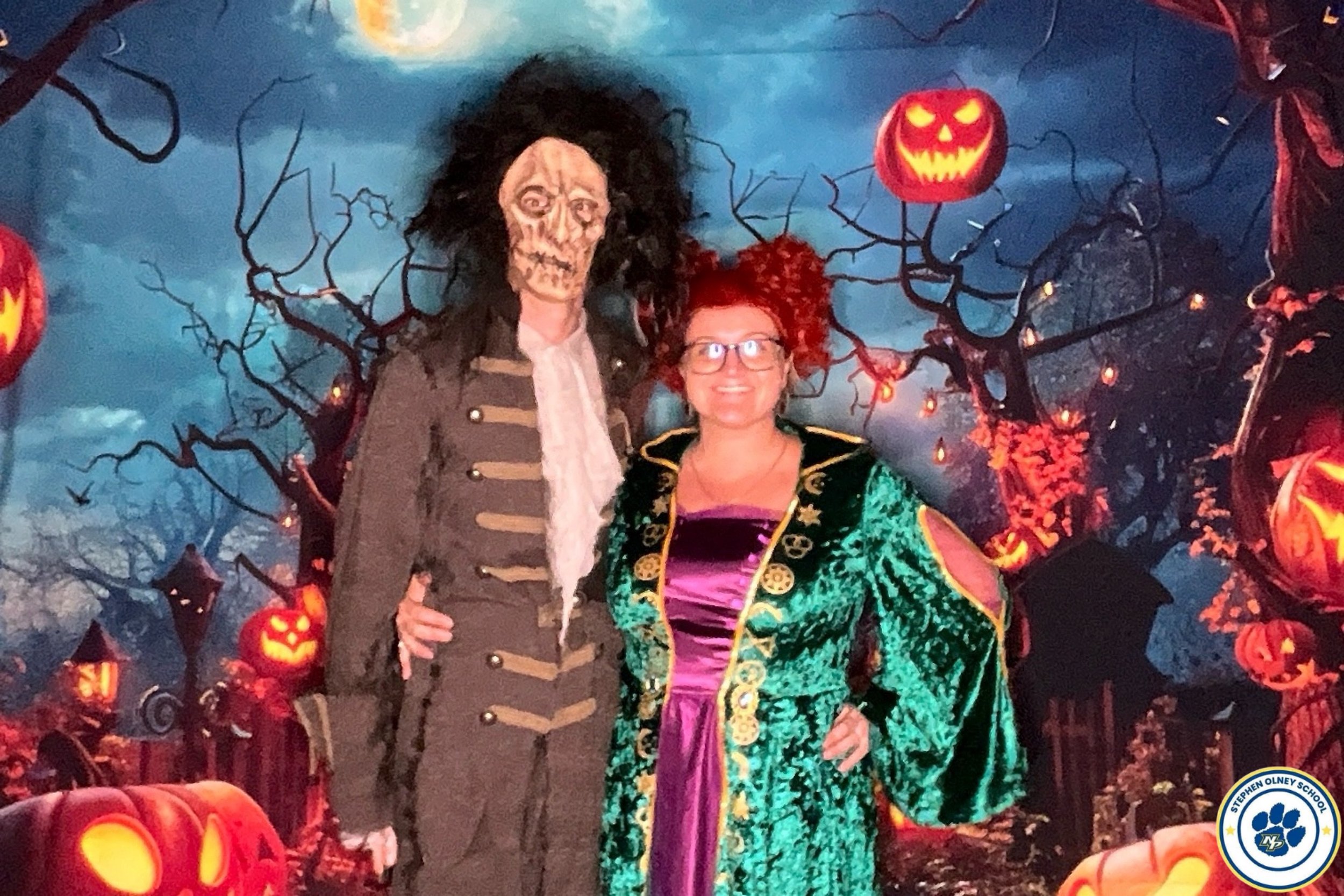 A woman dressed as a witch with red curly hair and glasses is standing next to a person in a skeleton costume with a zombie face mask. They are posing in front of a Halloween-themed background with carved pumpkins, spooky trees, and glowing lights.