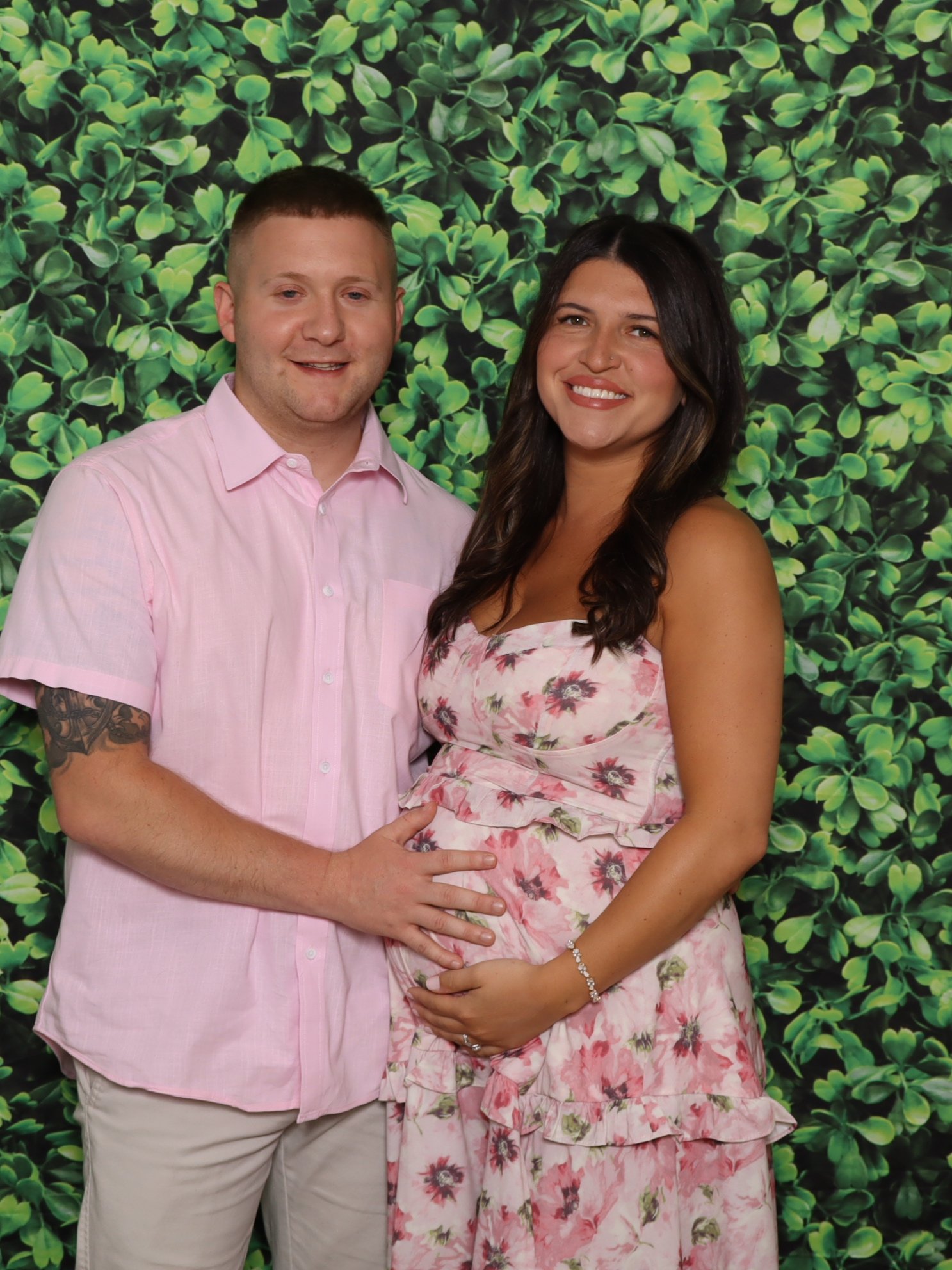 A smiling couple, a man in a light pink shirt and a woman in a floral dress, standing close together with their hands on the woman's pregnant belly, posed in front of a lush green leafy background.