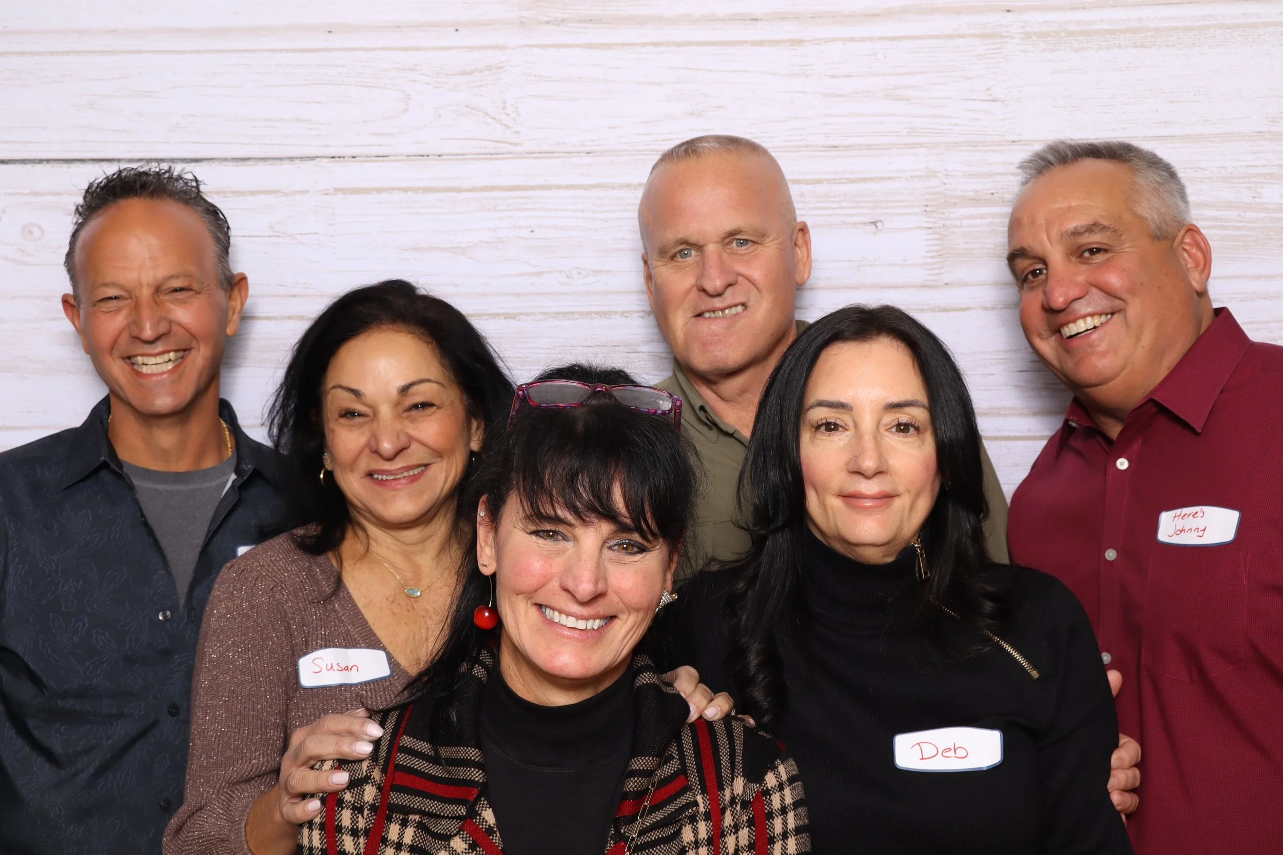 Group of seven middle-aged adults smiling, standing against a white wooden background, with some wearing name tags.