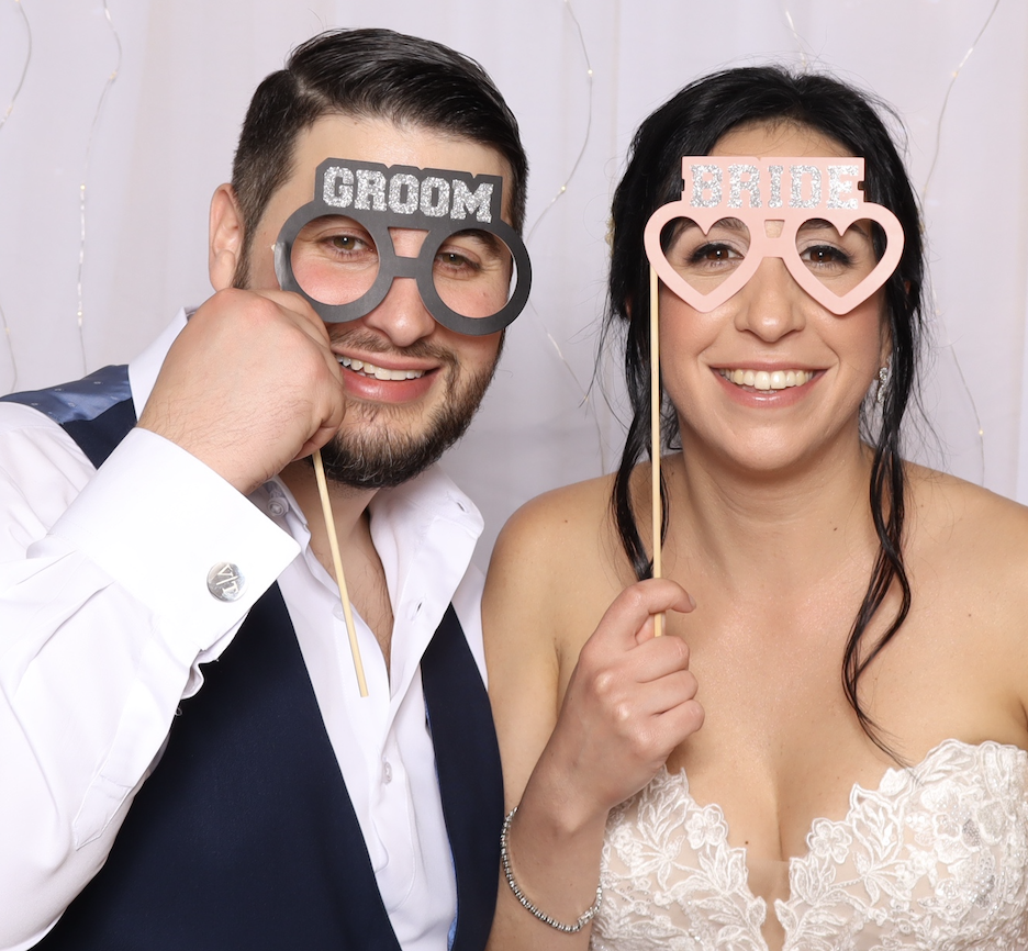 A happy couple at a wedding, holding fun photo props for the bride and groom, smiling at the camera.