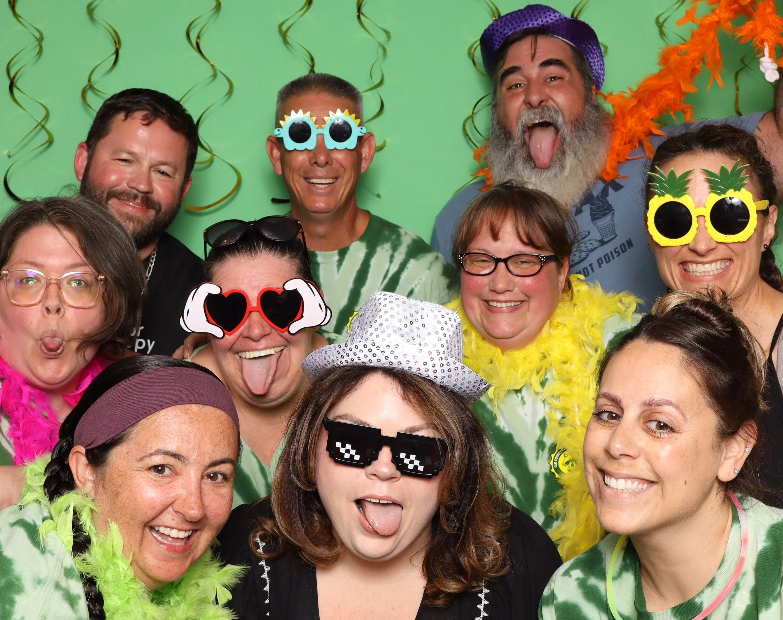 Group of people at a party wearing colorful costumes, glasses, and fun accessories, with a green backdrop and streamers.