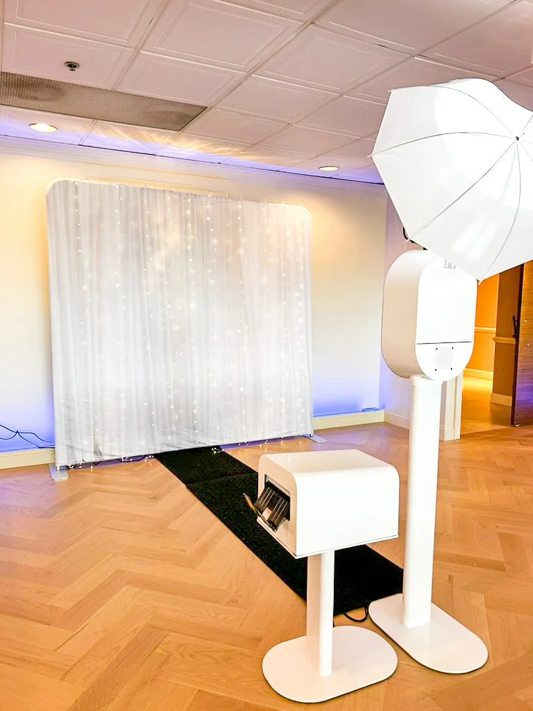 Photo of a photo booth with a black rug in front, white fabric backdrop with string lights, and a photobooth camera on a stand, all set up indoors on a wooden floor.