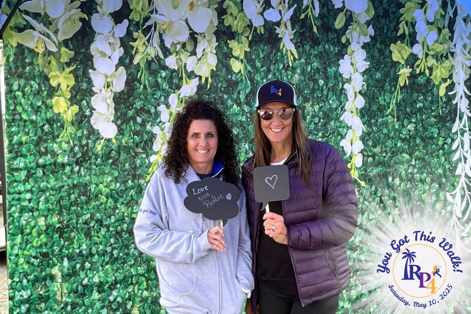 Two women smiling and holding signs, standing in front of a green leafy backdrop with white hanging flowers, at an event called 'You Got This Woe!' on Saturday, May 10, 2025, organized by RP4.