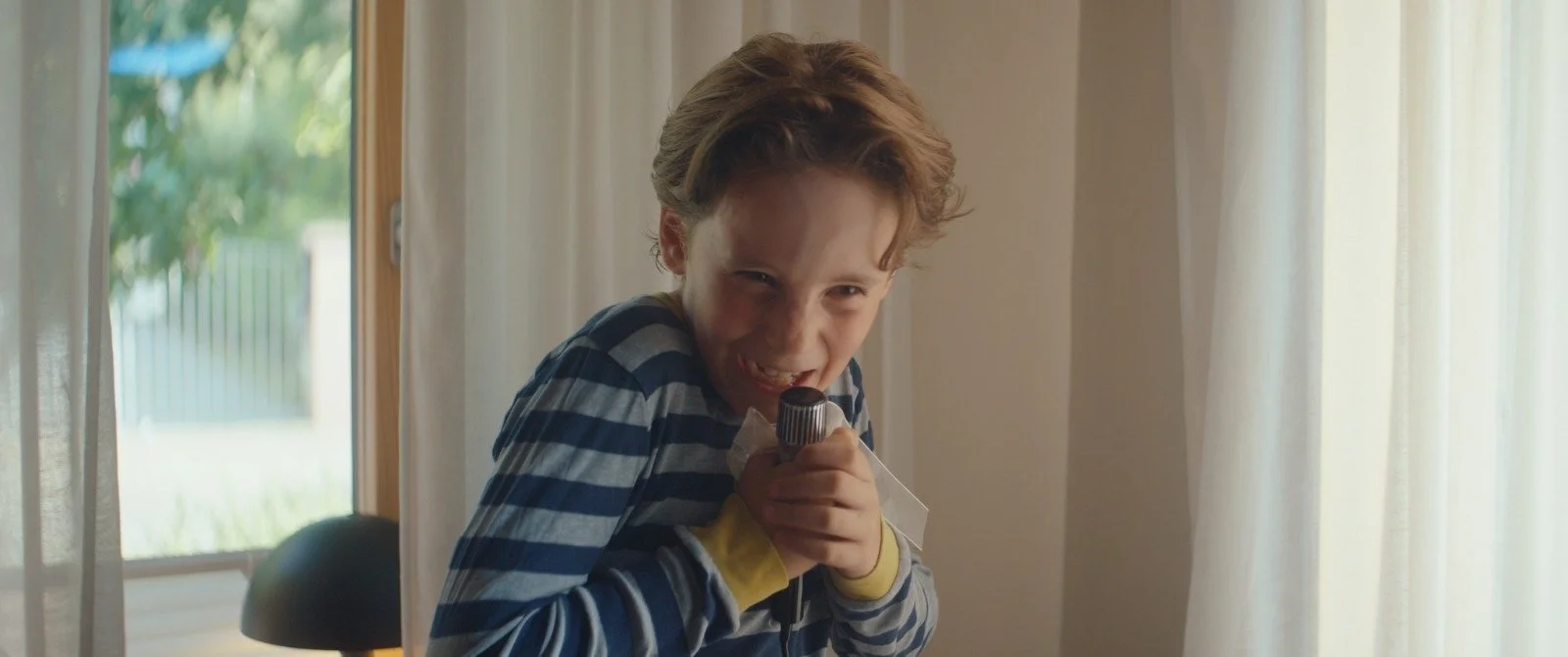 A young boy with curly hair wearing a striped shirt and yellow gloves is holding a microphone and pretending to sing.