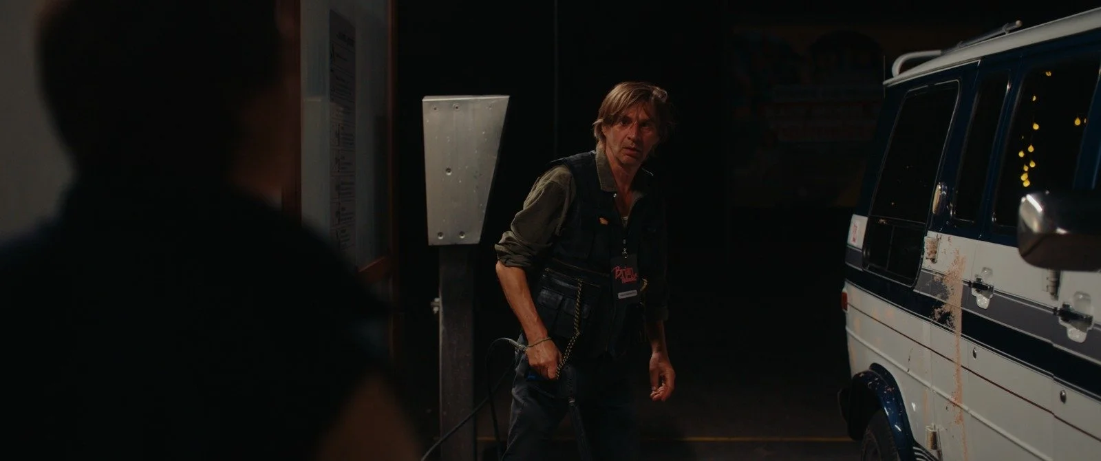 A man standing on a dark street at night, next to an ambulance with bloodstains, holding a leash, with other person partly visible in the foreground.