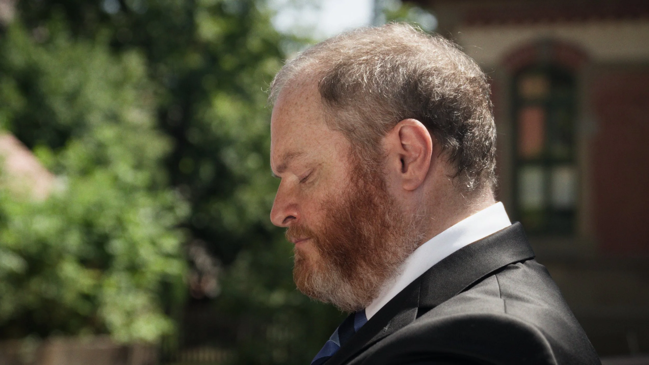 Profile of a man with a beard and short hair, dressed in a suit, outdoors during daytime.