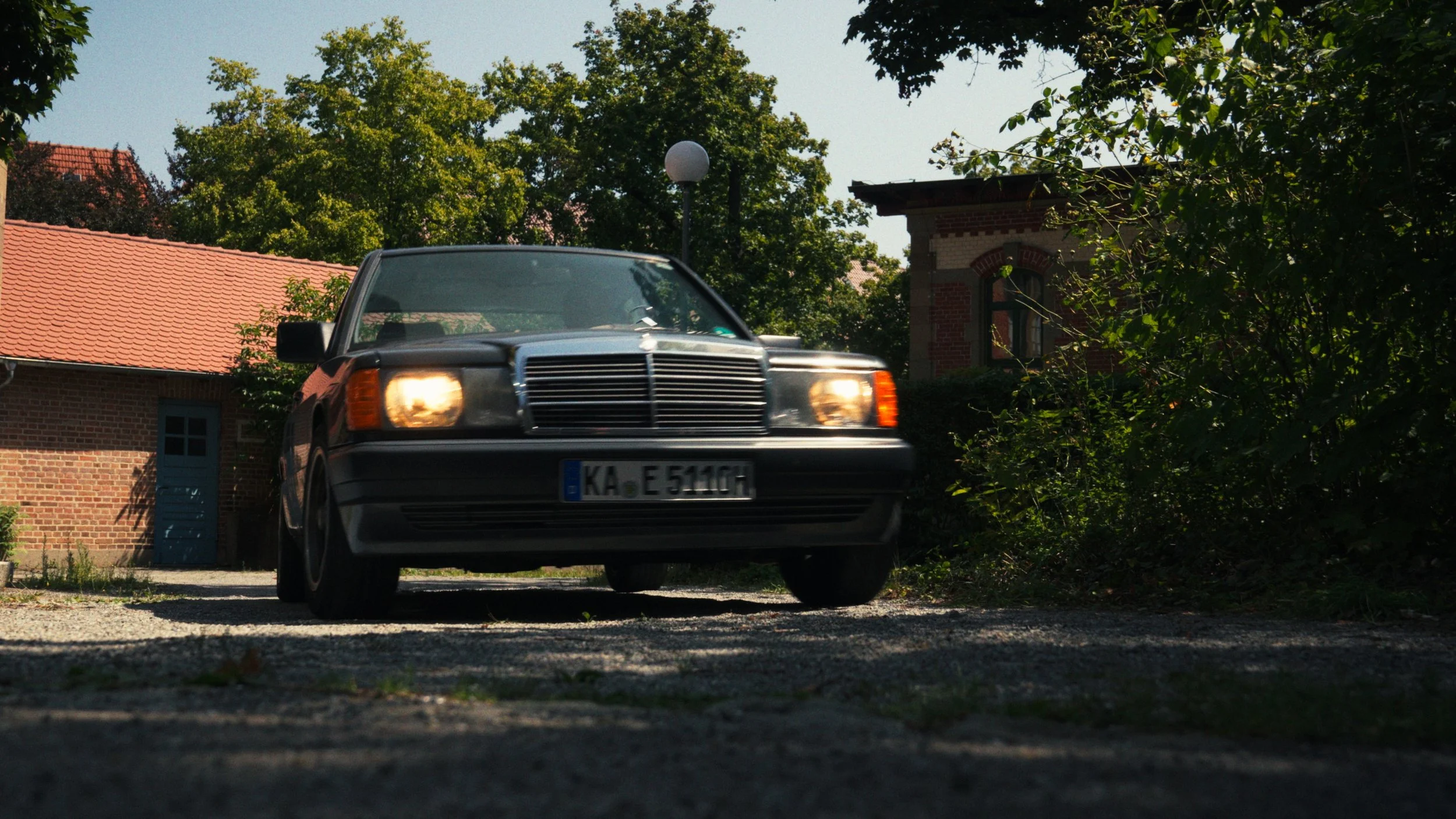 A black classic Mercedes-Benz car with its headlights on, parked on a gravel driveway in front of a brick house with a red tiled roof, surrounded by green trees and bushes.