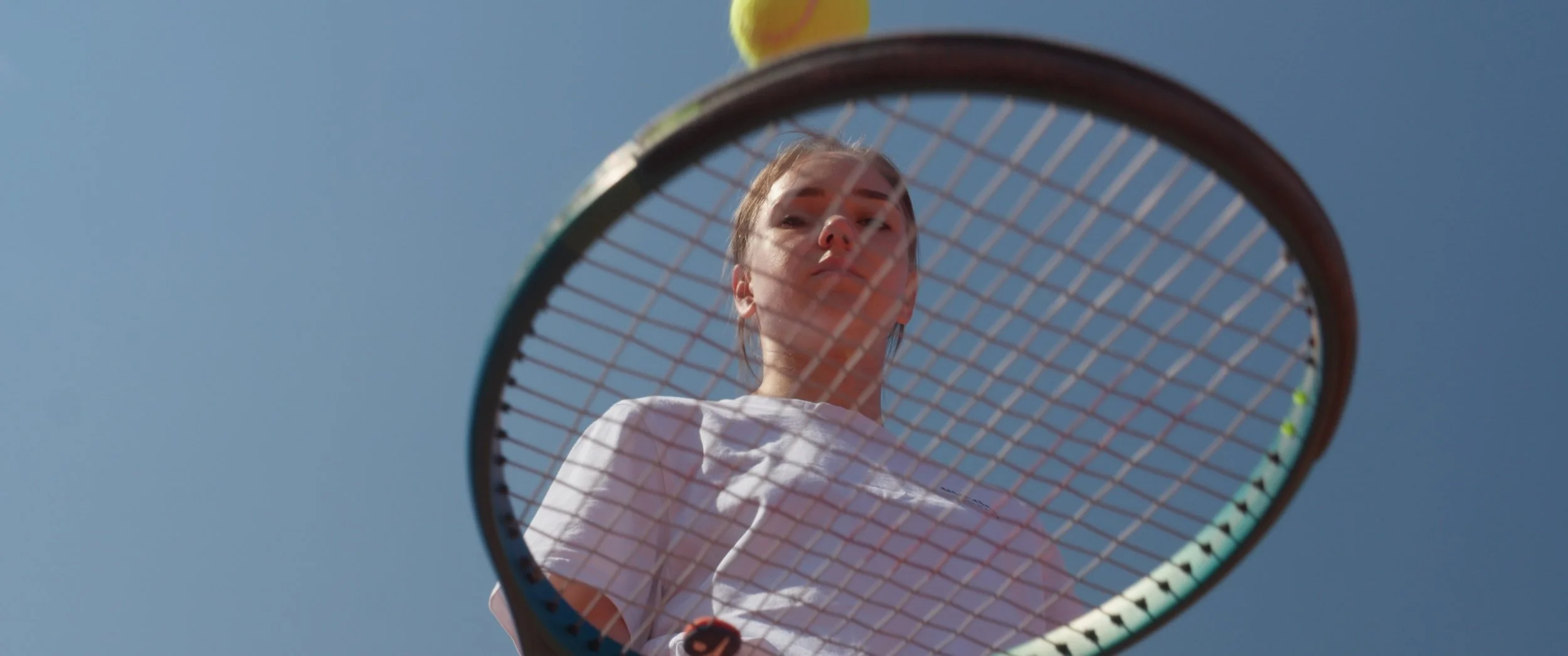 A woman with a tennis racket looking down at the camera, with a clear blue sky in the background.