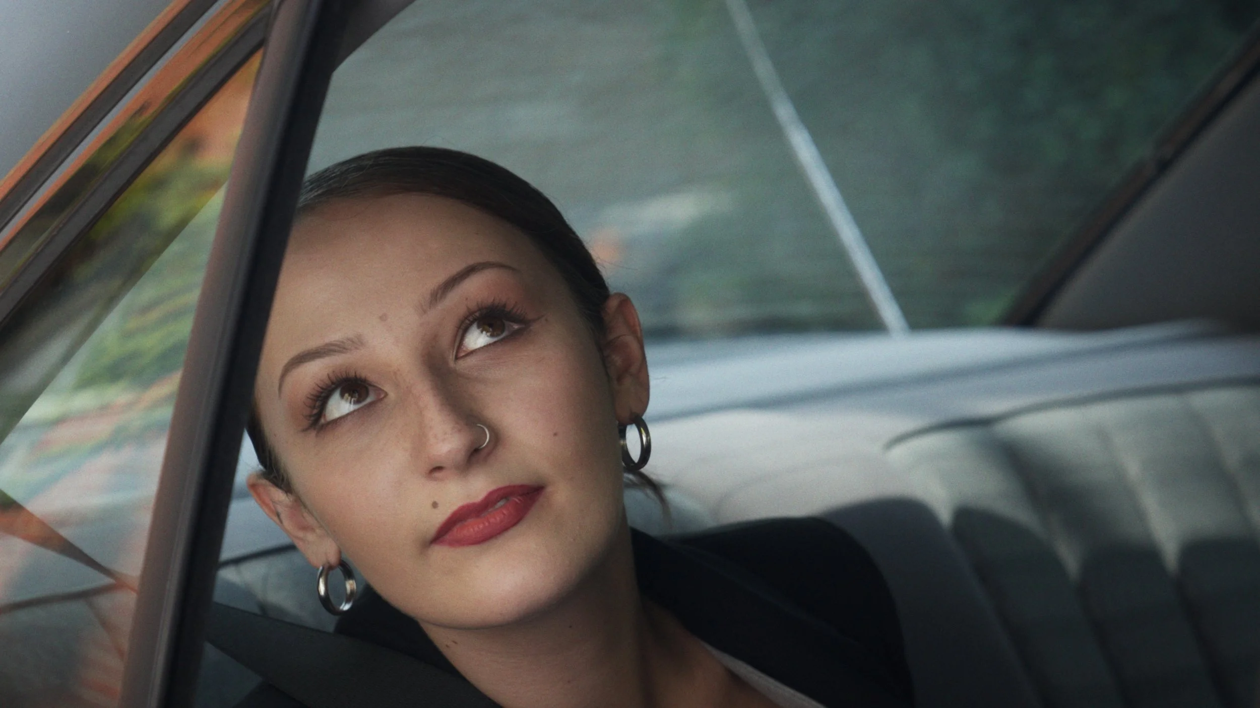 A young woman with dark hair and brown eyes looks up through a car window. She is wearing earrings and a nose ring, and has a calm expression.