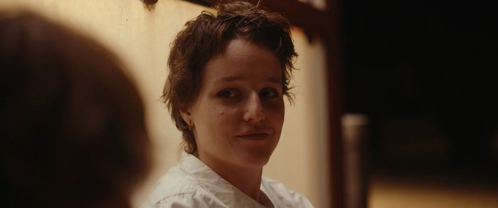 A woman with short, wavy hair and earrings, wearing a white shirt, looking sideways with a gentle smile in an indoor setting with warm lighting.
